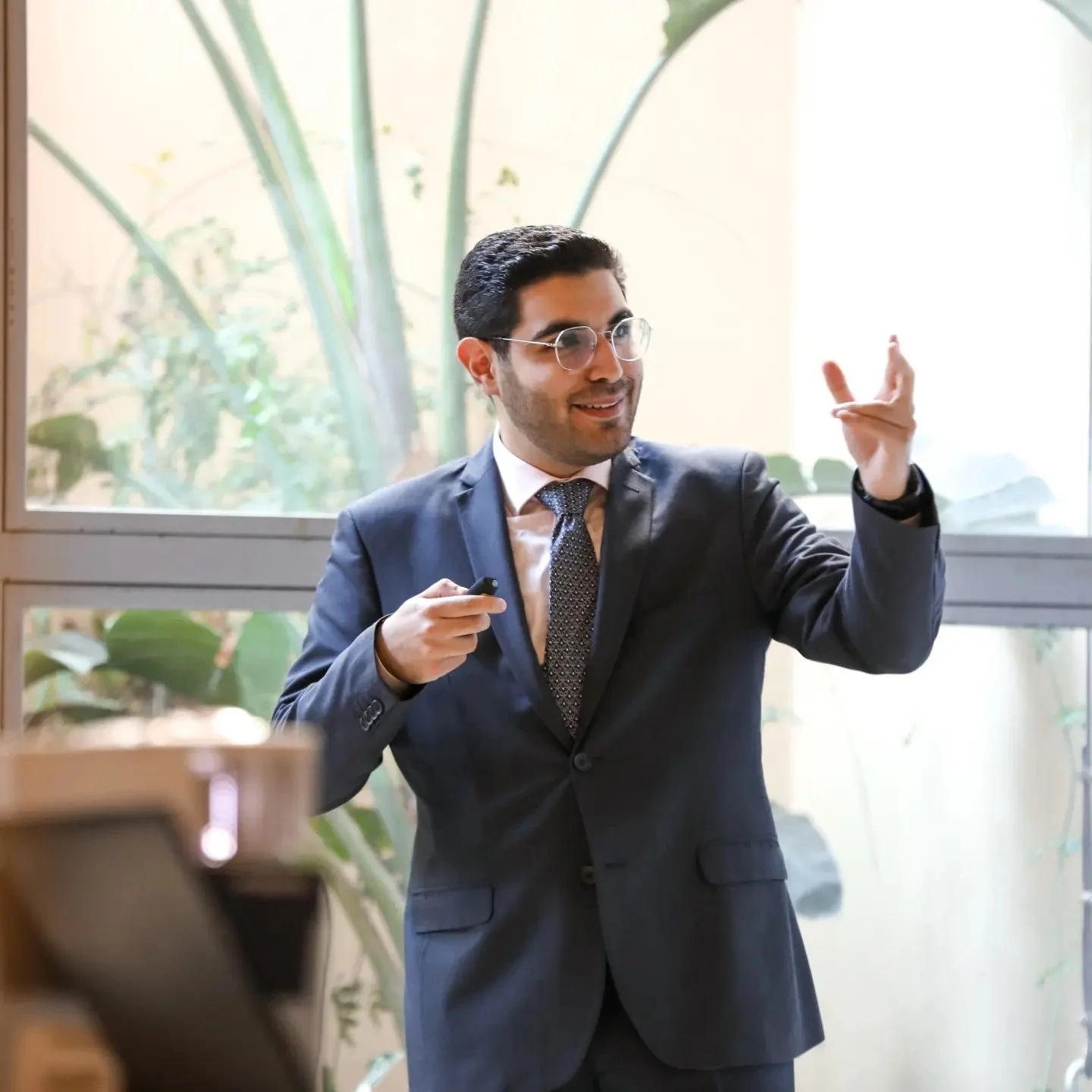 Man in a dark suit and glasses giving a presentation or speech in a room with large windows and tropical plants in the background.