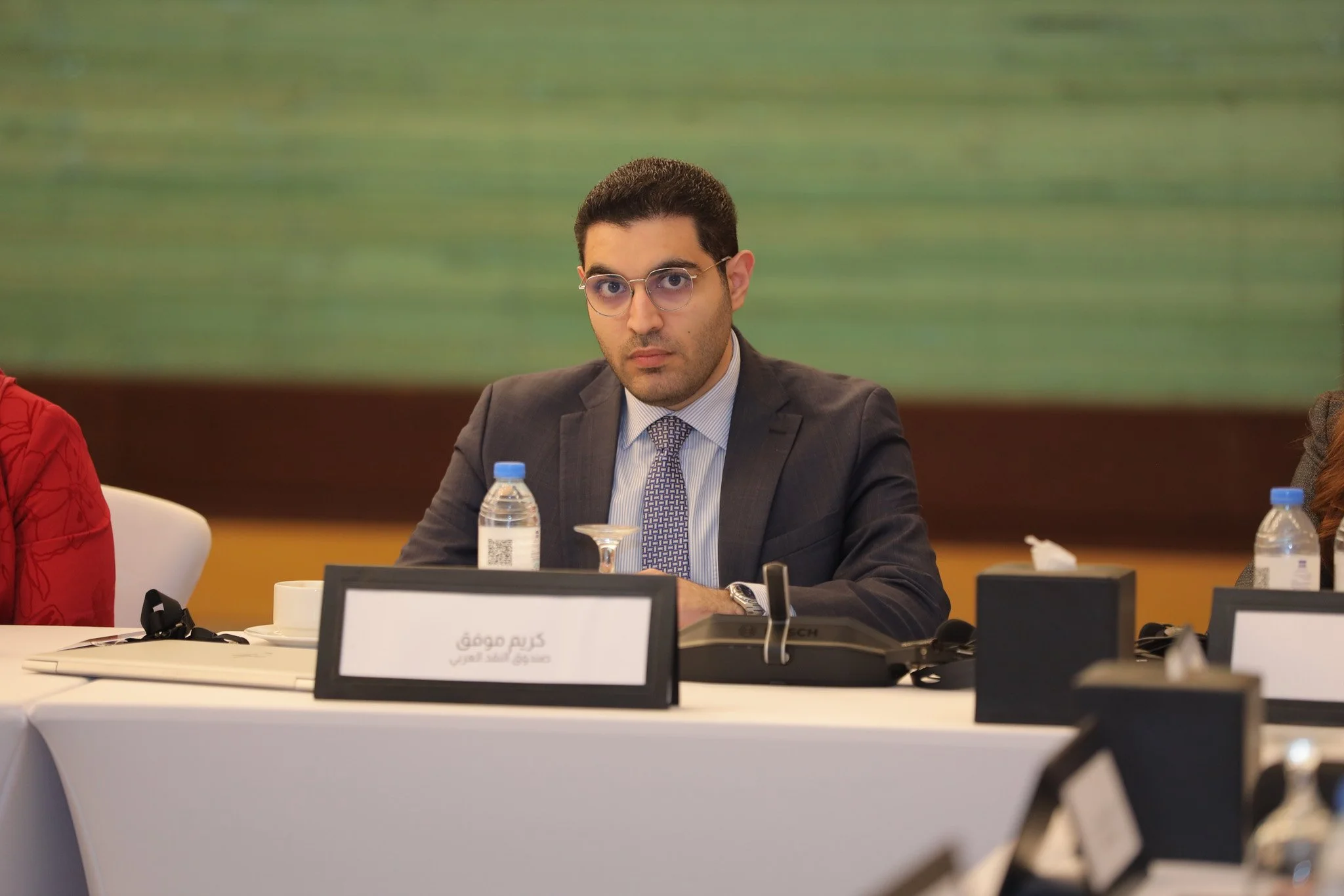 A man in a dark suit and glasses sitting at a conference table with water bottles, a nameplate, and a conference phone.