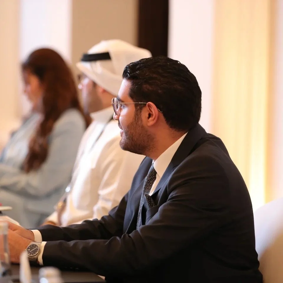 A man with black hair, glasses, and a beard, dressed in a black suit and tie, sitting at a conference table, smiling during a meeting.