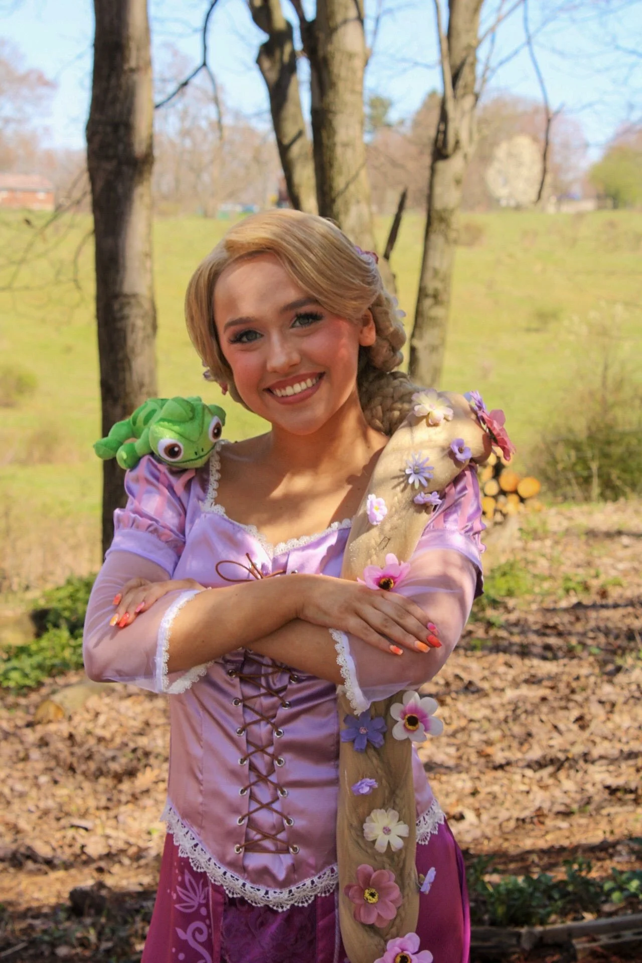 Rapunzel is smiling and holding a wooden staff covered in flowers. She has a plush turtle on her shoulder and is outdoors in a wooded area.