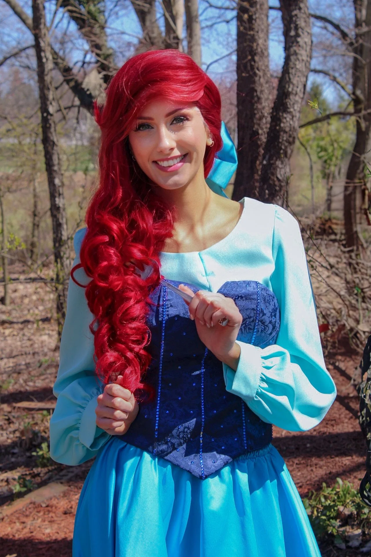 Ariel with long red curly hair, wearing a blue and white princess dress, standing outdoors in a wooded area with trees and sunlight.