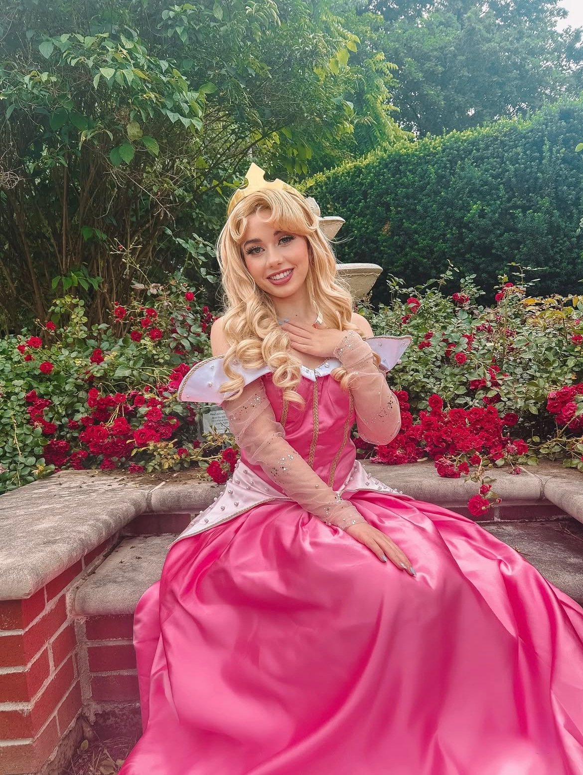 Sleeping Beauty or Princess Aurora in a pink gown sitting on a stone bench in a garden with pink and red flowers and green bushes.