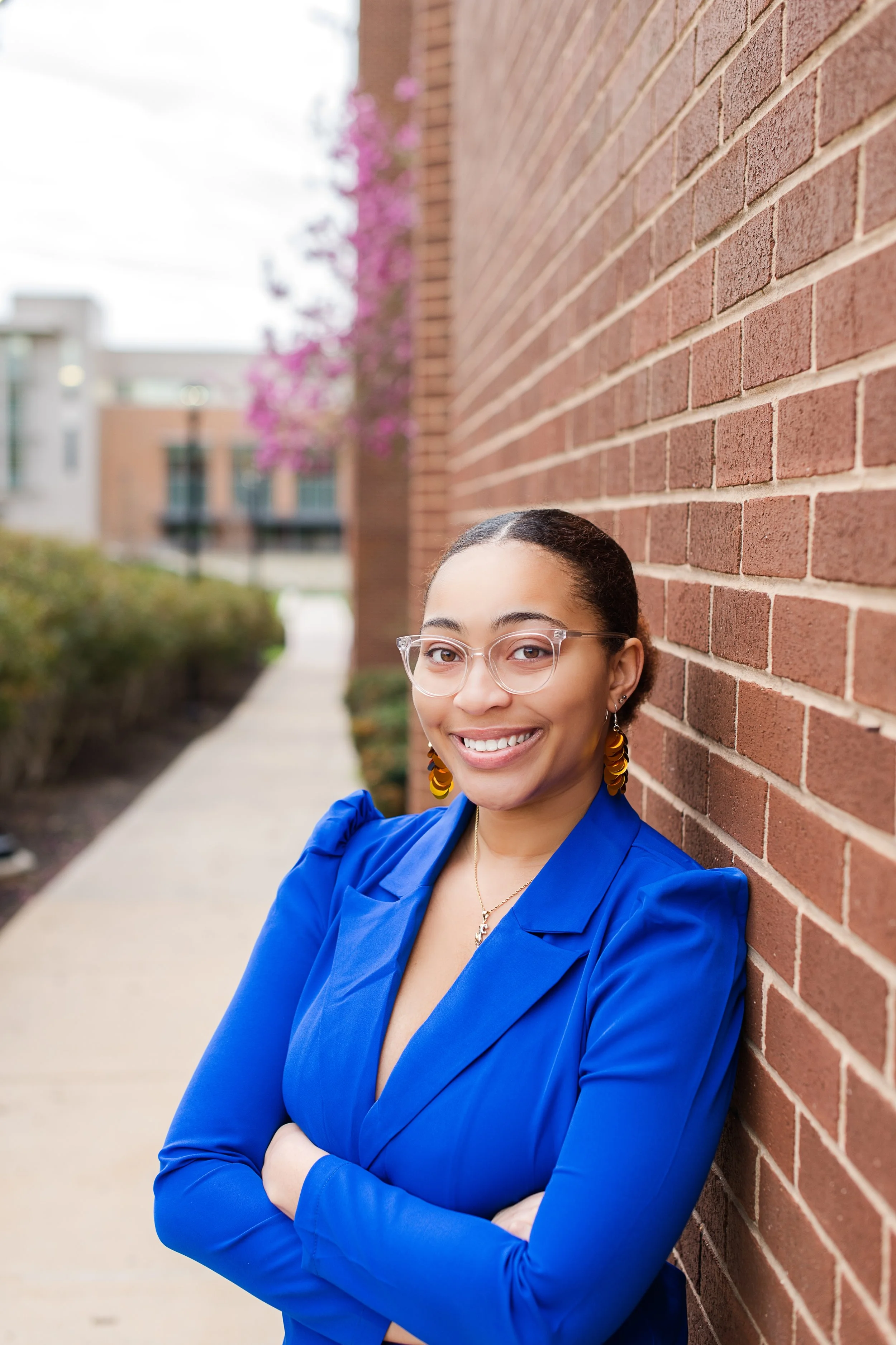 Smiling woman with glasses and earrings leaning against a brick wall outside in an urban setting.
