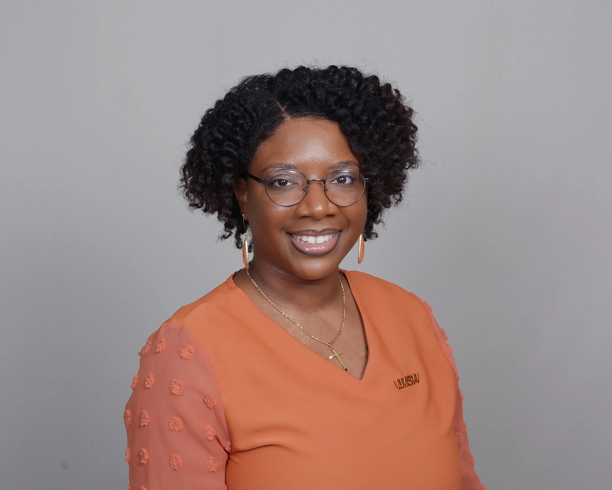 A woman with curly black hair and glasses, wearing earrings, a necklace with a cross, and an orange top with textured sleeves, standing against a gray background.