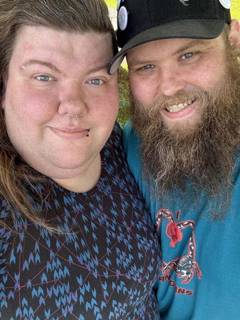 Close-up of a woman and a man smiling, outdoors. The woman has facial piercings and long brown hair. The man has a thick beard, mustache, and wears a baseball cap.