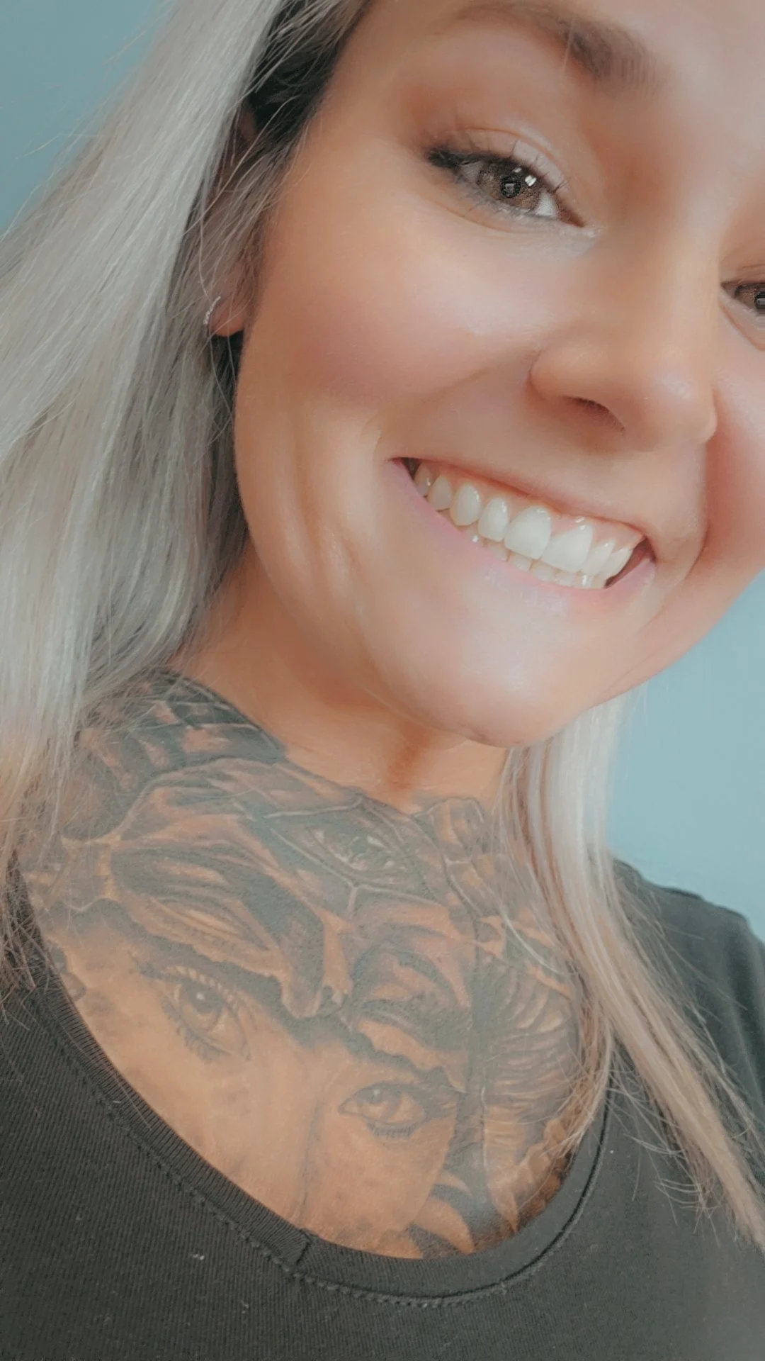 Close-up of a smiling woman with blonde hair, a tattoo of a tiger with two human eyes on her chest, and wearing a black top. She has makeup on and appears happy.