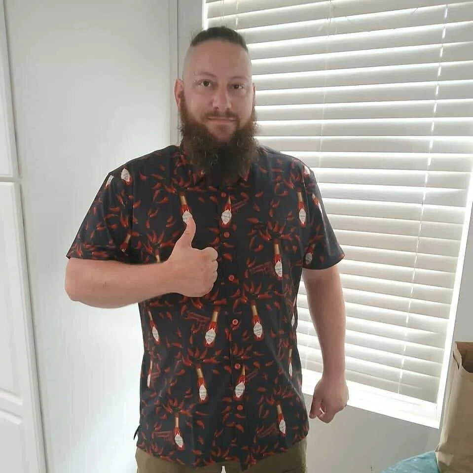A man with a beard giving a thumbs-up gesture, wearing a dark short-sleeved shirt with a Christmas bottle pattern, standing indoors next to a window with white blinds.