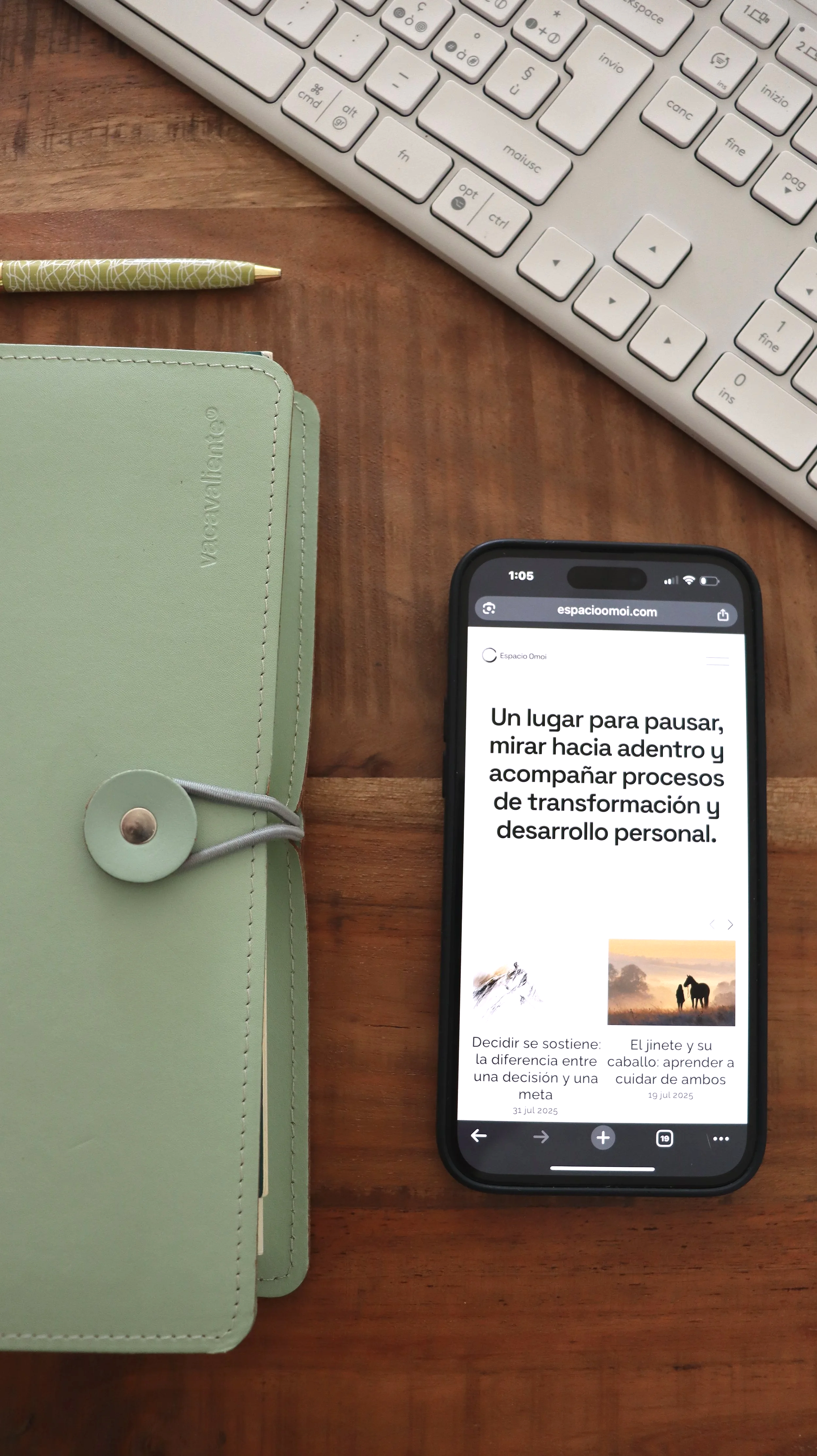 Un teclado, un cuaderno verde, una pluma y un teléfono móvil en una mesa de madera. La pantalla del teléfono muestra un sitio web con texto en español que describe un espacio para la transformación y el desarrollo personal.
