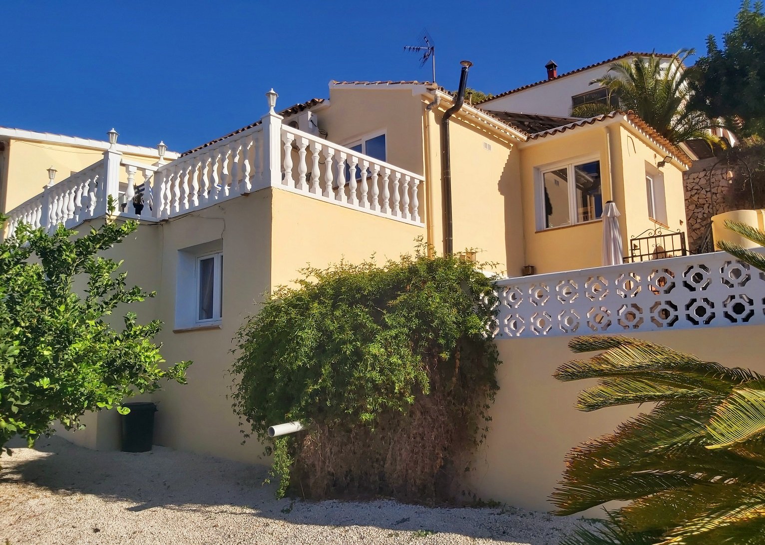 Mediterranean-style house with beige walls, white balustrade, garden plants, and blue sky.
