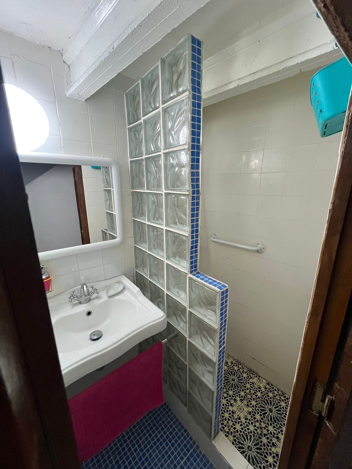 Small bathroom with glass block shower wall, white sink with faucet, mirror, blue and white tile floor, pink towel, and shower area with grab bar.