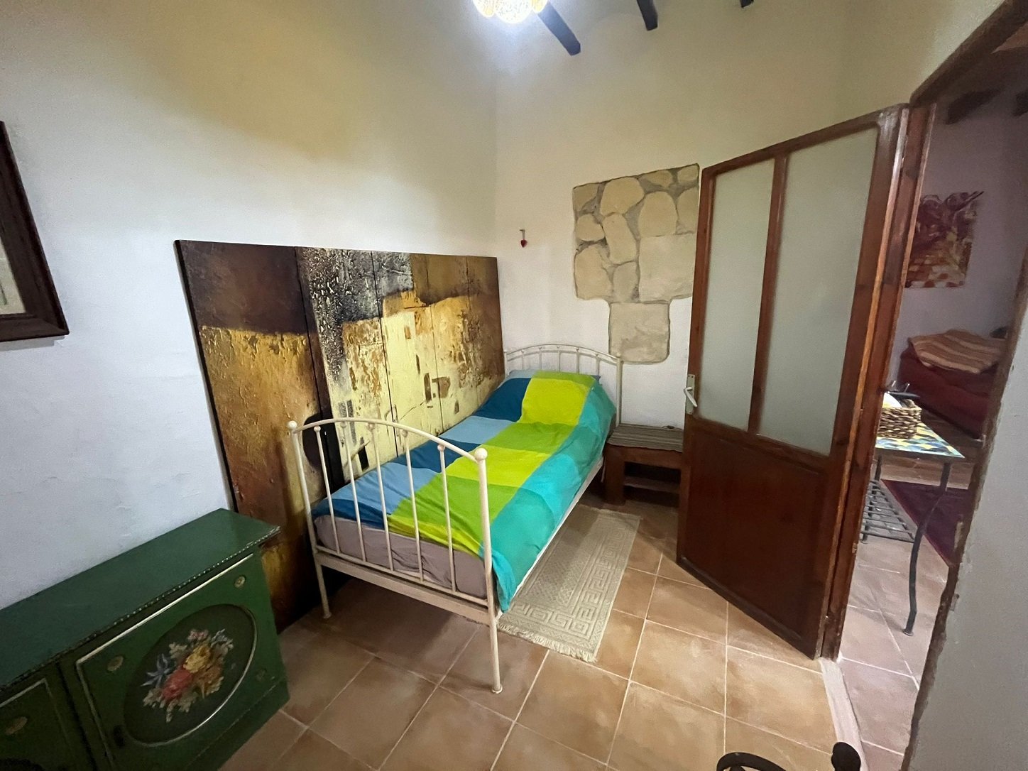 A small room with a single bed featuring a colorful duvet, wall art, a decorative wooden cabinet, and a glass-paned door.