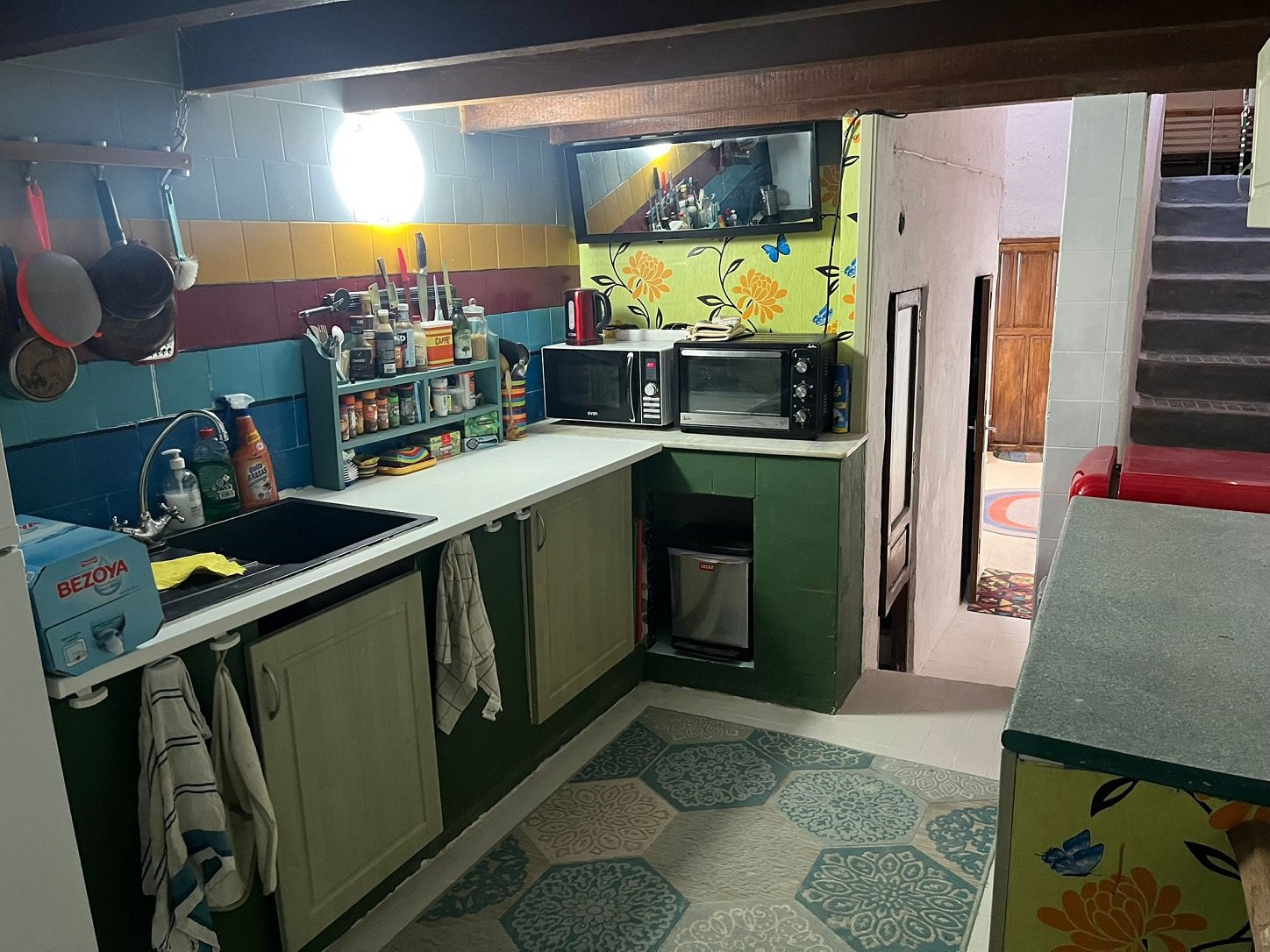 Colorful kitchen with tiled walls, wall-mounted pans, countertop with sink and dish rack, assorted kitchenware, microwave, oven, and vibrant floral wallpaper.
