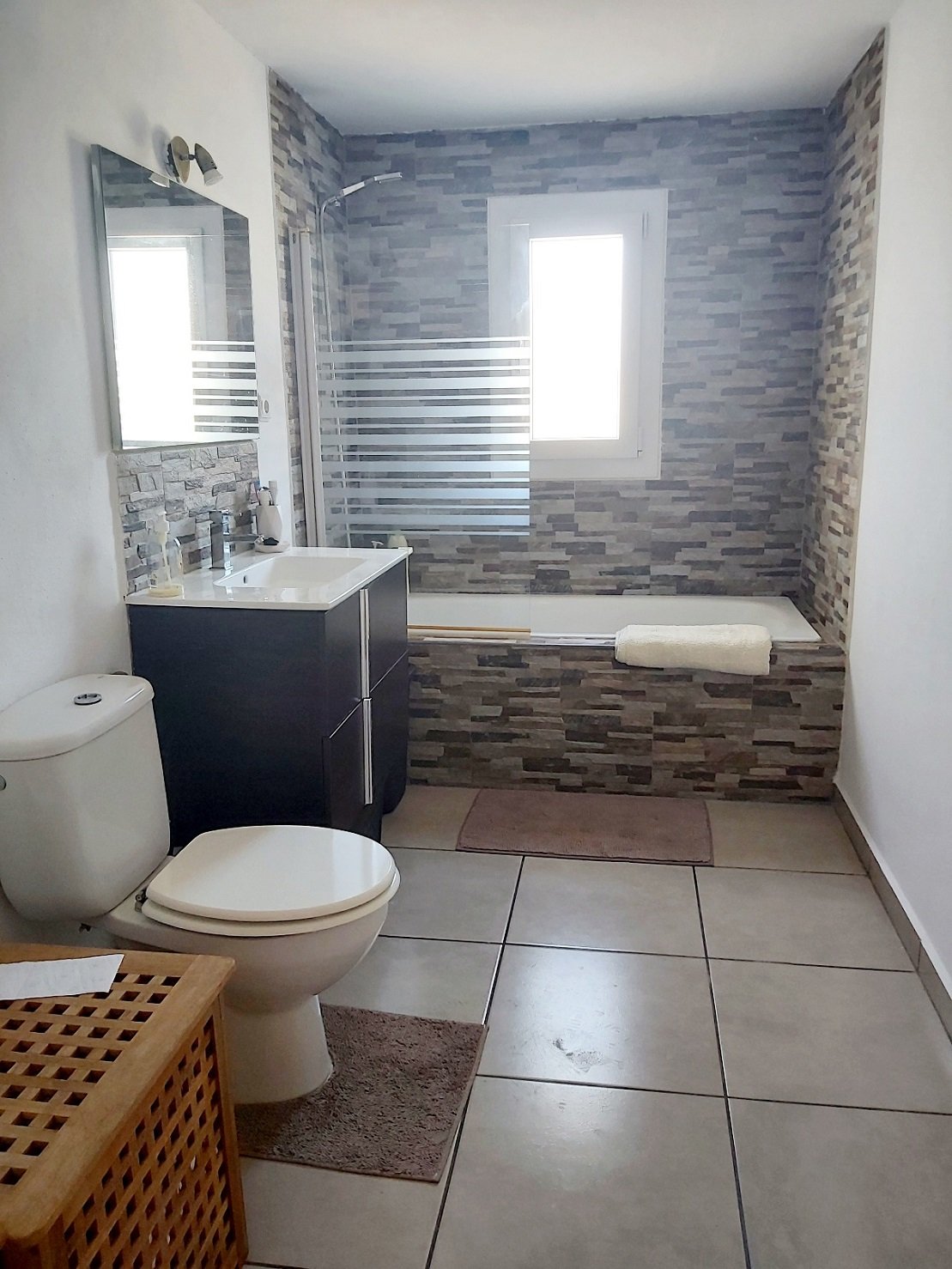 Modern bathroom with tiled walls, bathtub with glass screen, white toilet, sink with mirror, and window.