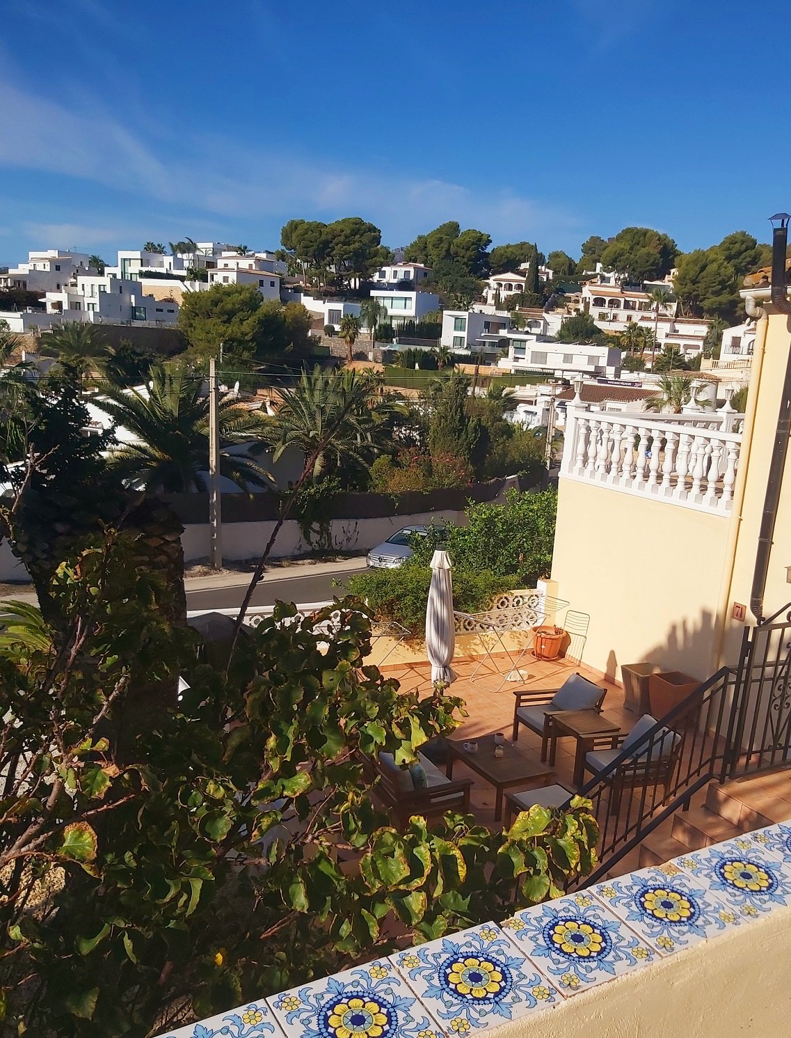 View of a Mediterranean hillside neighborhood with white houses amidst greenery, featuring a foreground patio with outdoor furniture and decorative tiles.