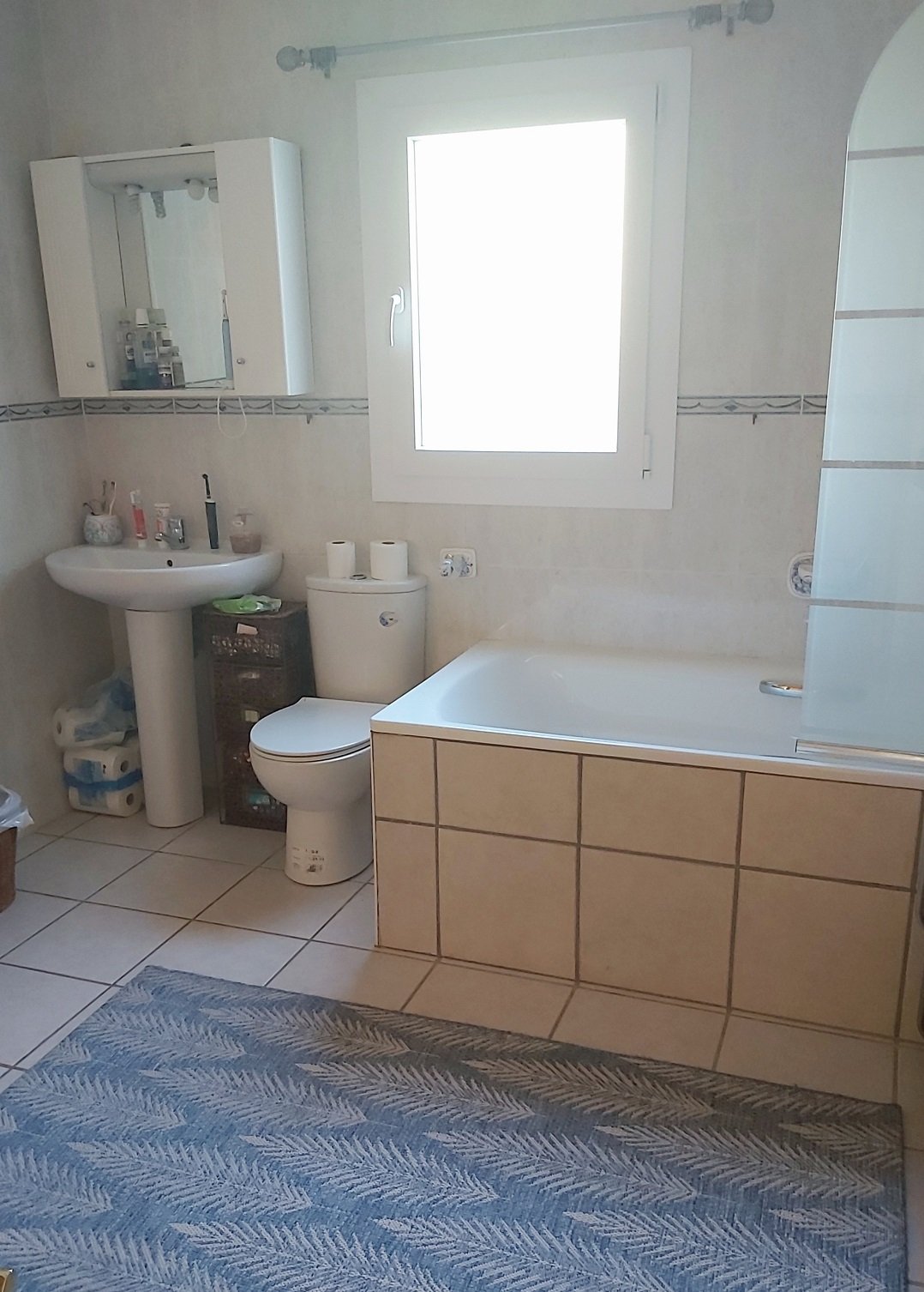 Bathroom with tiled floor, bathtub, toilet, sink, mirror cabinet, window, and toiletries.