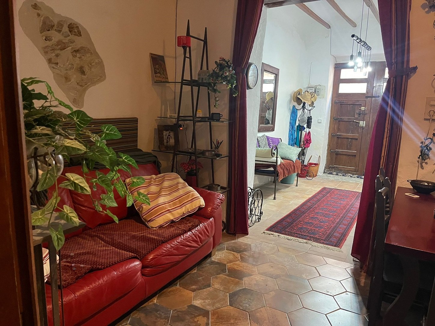Cozy living space with red couch, decorative pillows, potted plants, and tiled floor. A bookshelf and wall art are visible. Entry hallway has a bench, hats, colorful scarf, and wooden door with glass panels. Red patterned rug leads to dining area wit