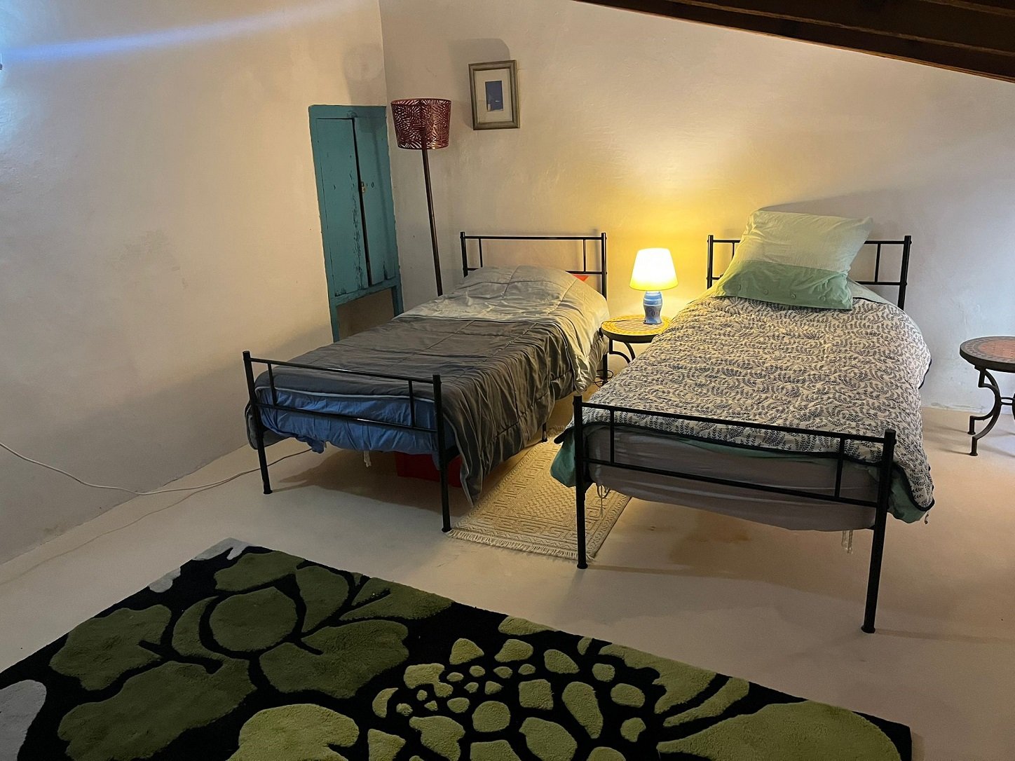 Cozy bedroom with two single beds, patterned bedding, a small bedside lamp, a decorative area rug, colorful cushion, and rustic decor. Warm ambient lighting with a small framed picture on the wall.