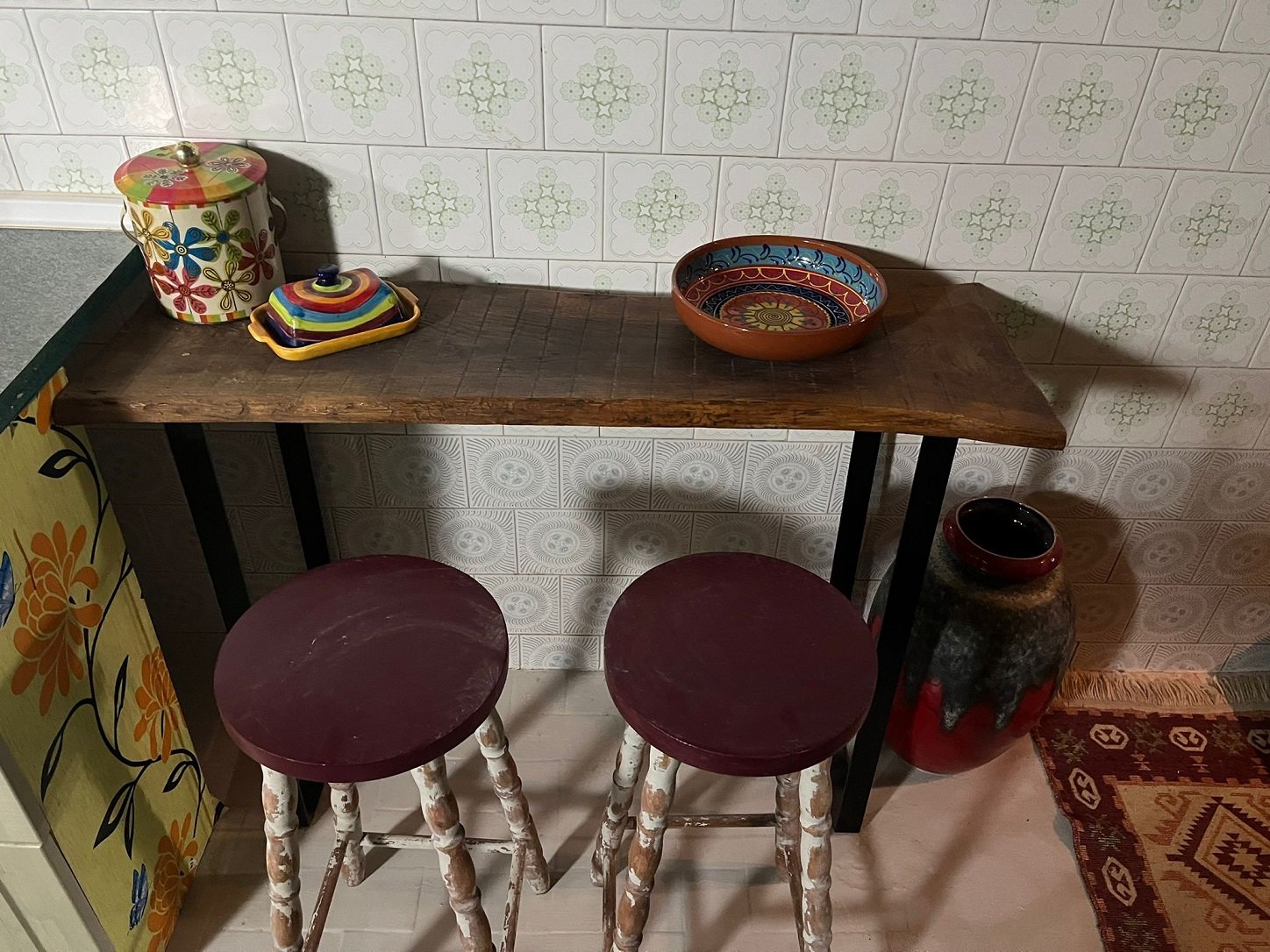 Vintage kitchen counter with colorful ceramic containers, two red stools, decorative bowl, jar, and patterned tiles.
