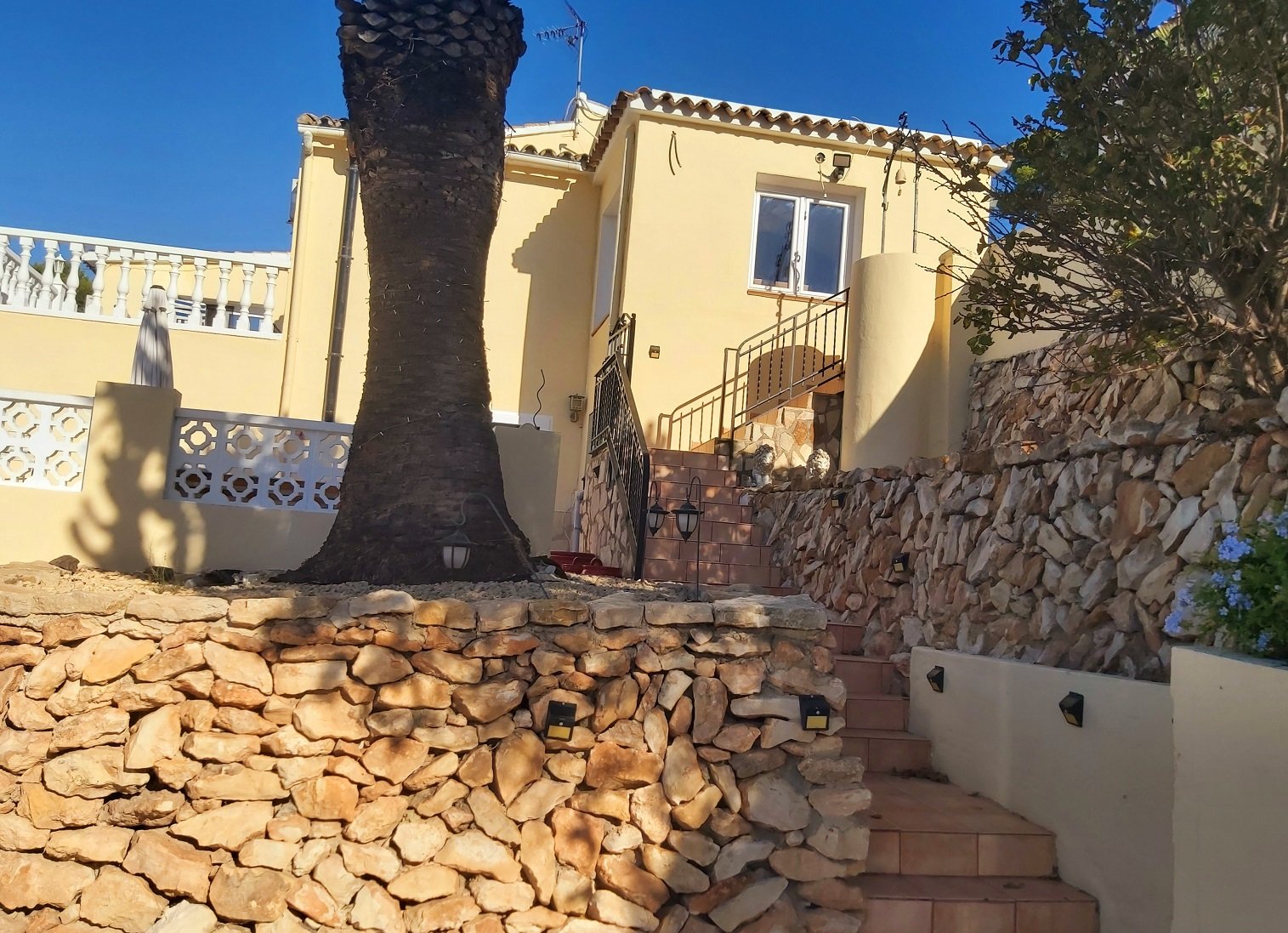 Mediterranean house with stone walls and a large palm tree in the foreground, featuring an outdoor staircase and decorative railings.
