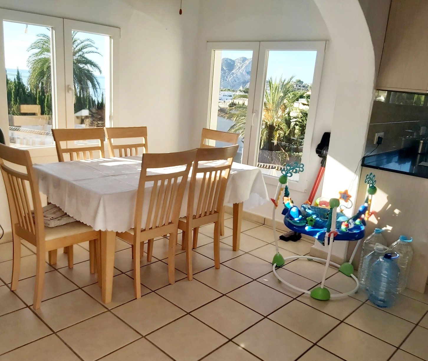 Dining area with wooden table and chairs, indoor scenery showing bright windows revealing palm trees and mountains outside, a baby activity bouncer in the corner, and large water bottles on the floor.
