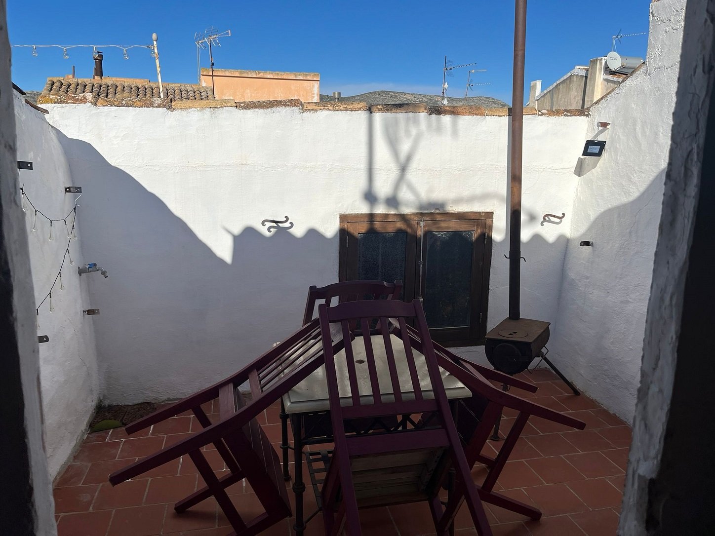 Rooftop terrace with folded chairs on a table, white walls, string lights, and metal chimney pipe.