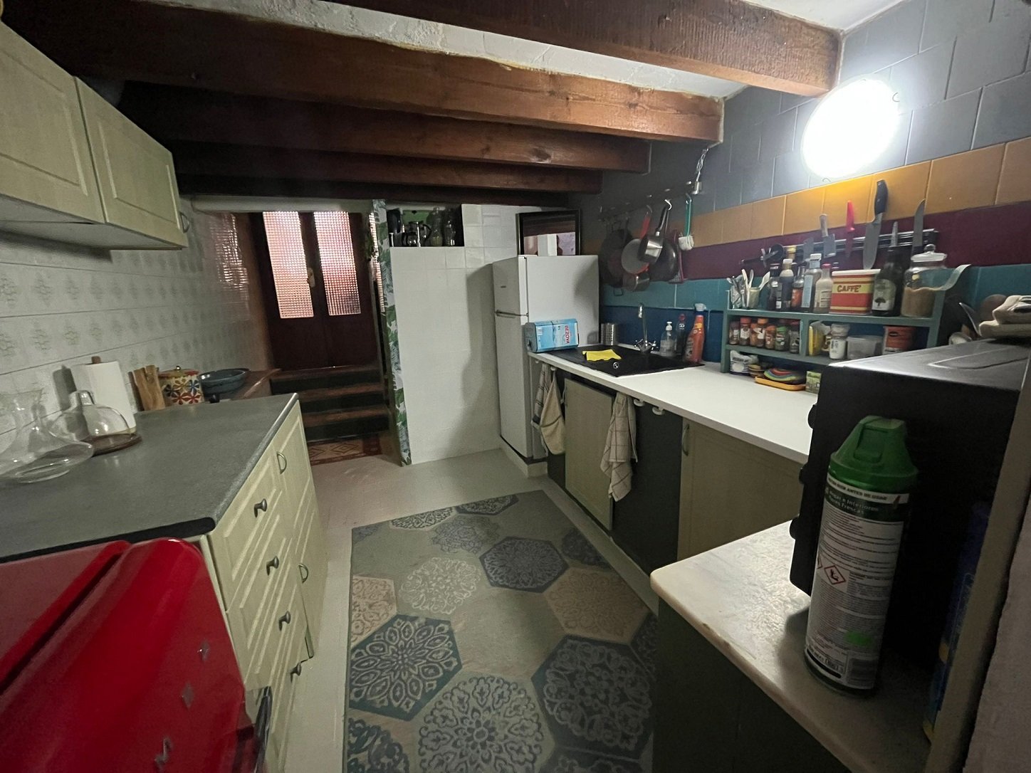 Cozy kitchen with wooden beams, patterned rugs, red fridge, and shelves with utensils and spices.
