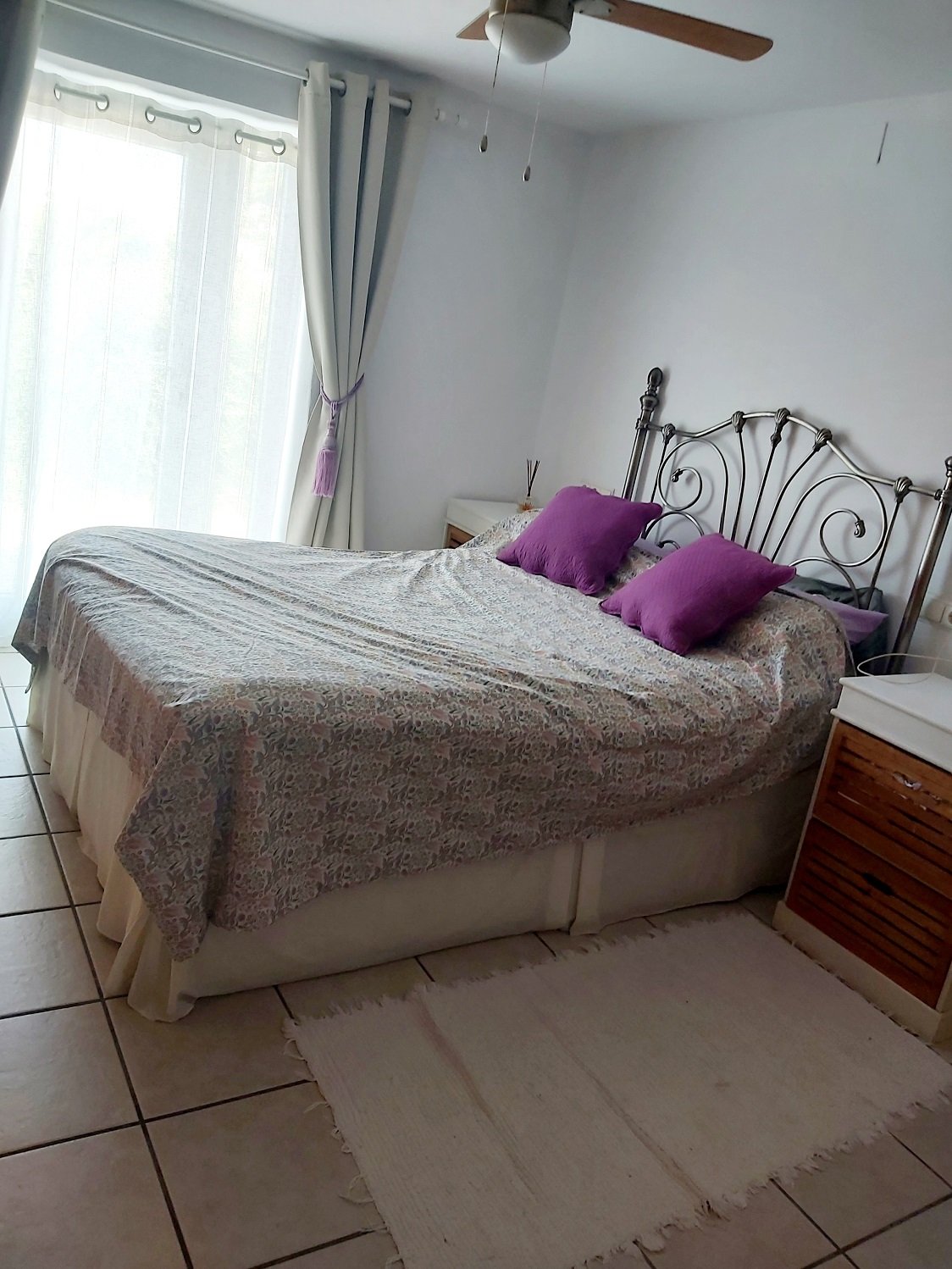 Bedroom with a large bed featuring a patterned bedspread and purple pillows, an iron headboard, a ceiling fan, white curtains, a wooden nightstand, and a light-colored rug on tiled floor.