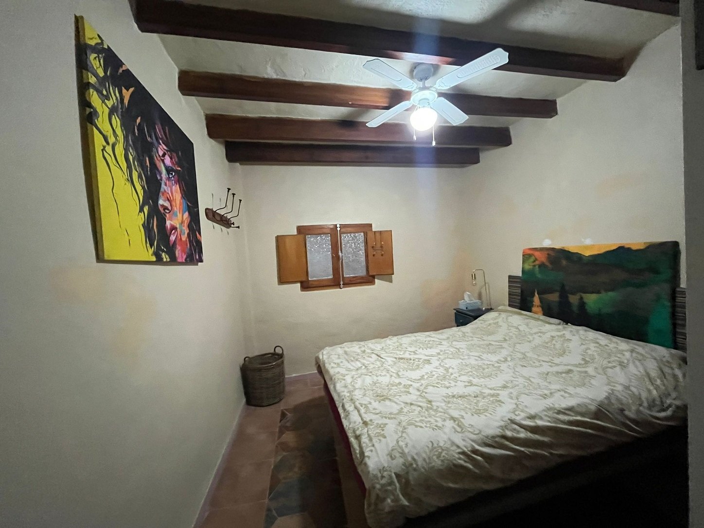 Small bedroom with a bed, ceiling fan, wooden beams, framed artwork on the wall, and a small window.