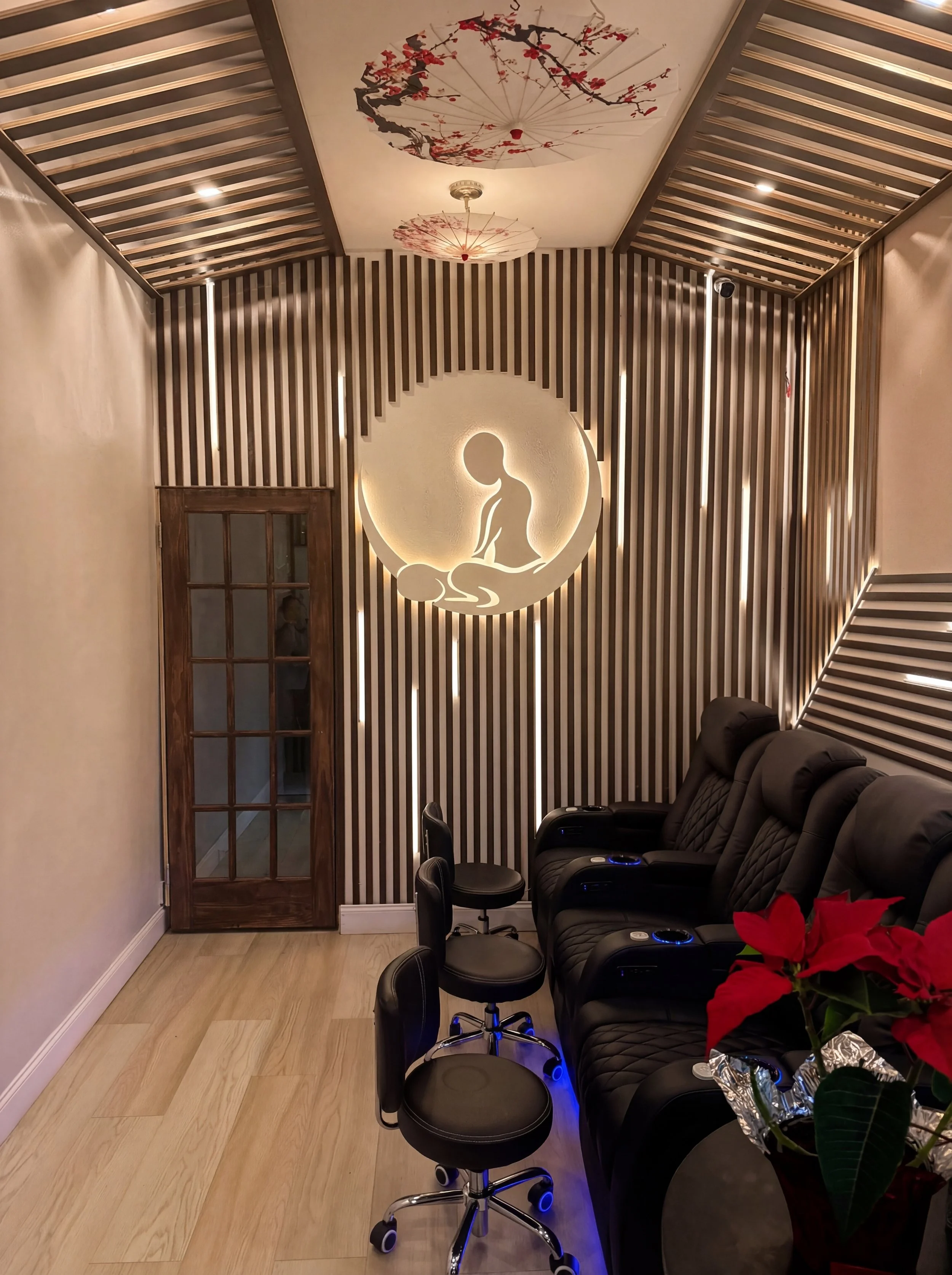 A spa or massage therapy room with four black leather massage chairs, a small black stool, a poinsettia plant, and a wall with wooden slats illuminated behind a circular cutout depicting a massage scene of a seated woman. The ceiling has decorative umbrellas with cherry blossom designs.