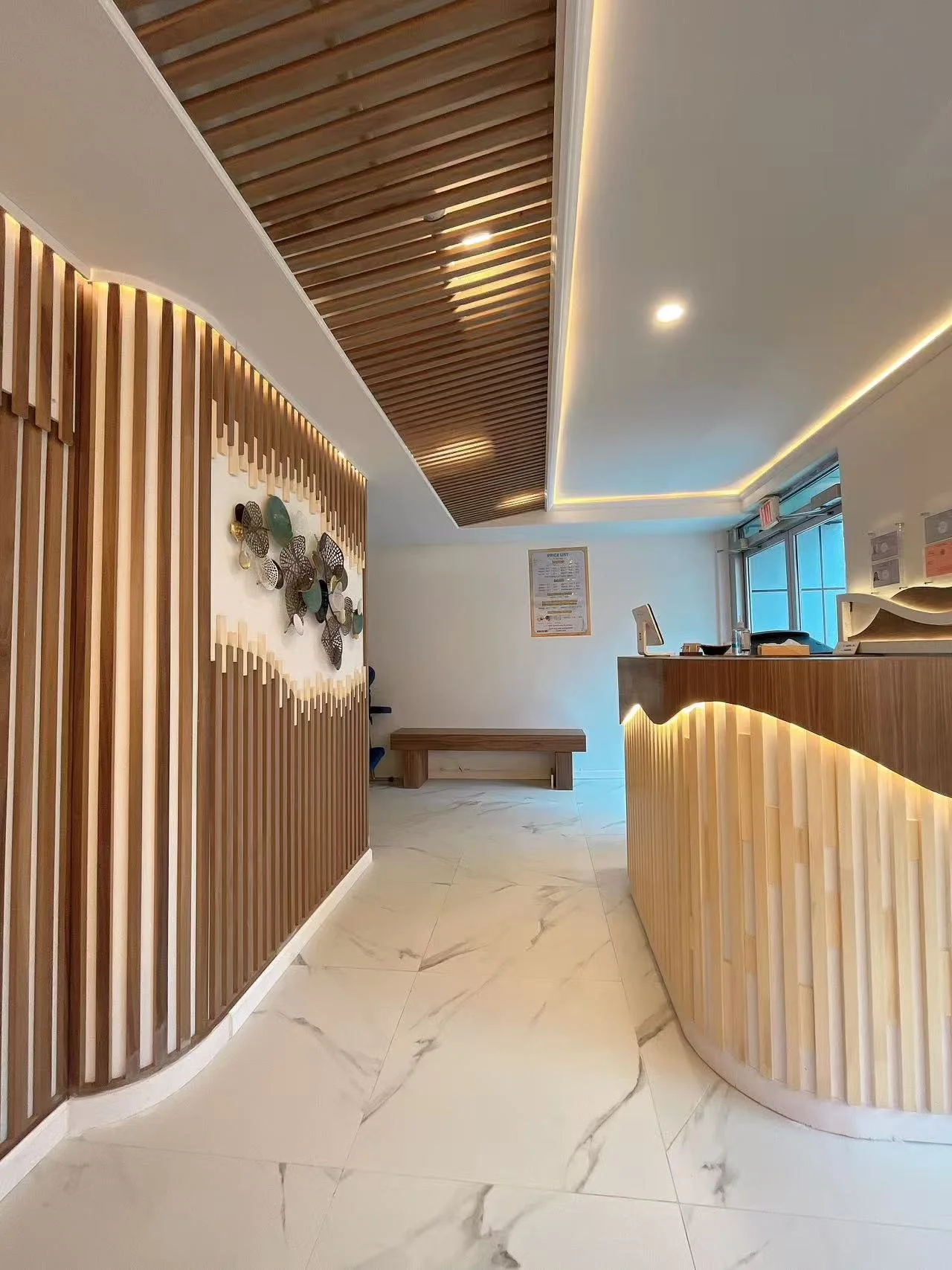 Modern reception area with curved wooden reception desk, decorative wall art, wooden slat ceiling, marble floor, and a bench in the background.