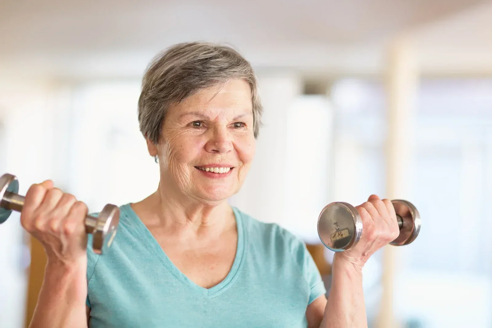 An older woman in a light blue shirt smiles while lifting dumbbells indoors. She appears cheerful and focused, embodying strength and vitality.