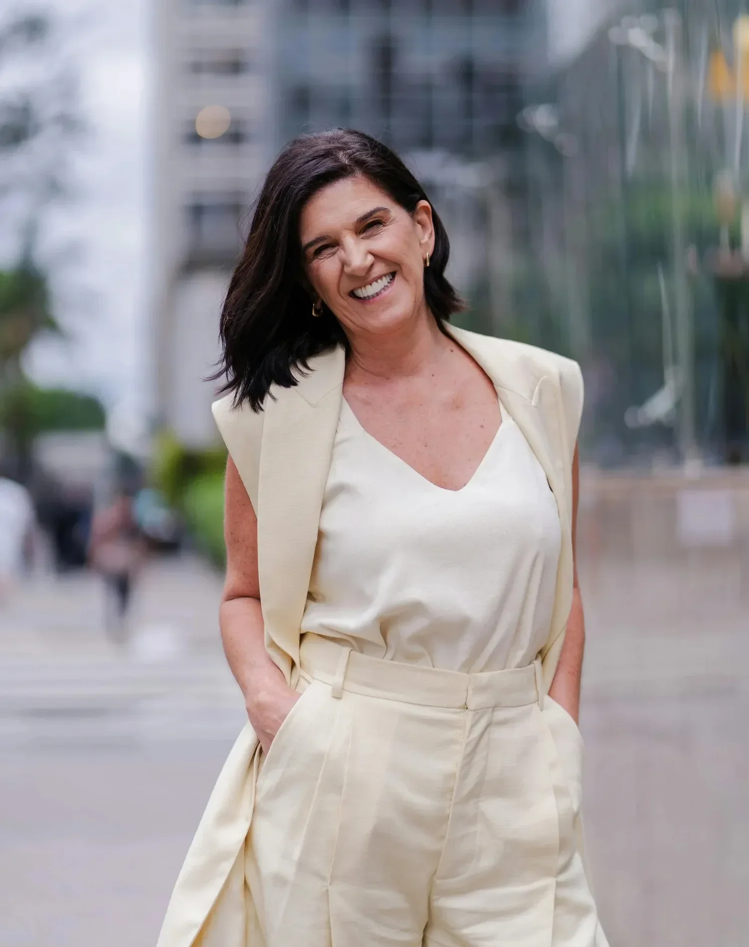 A woman in a cream outfit smiles confidently outdoors. She stands with hands in pockets on an urban street, conveying a professional yet relaxed vibe.