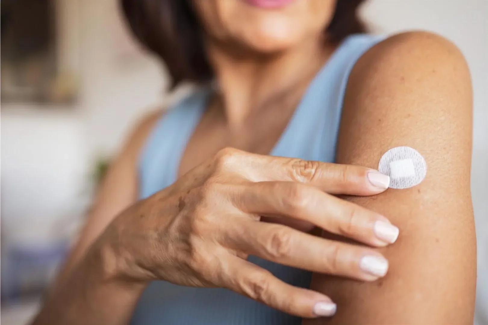 A person in a sleeveless top gently touches a small circular adhesive patch on their upper arm, suggesting health monitoring or medication delivery.
