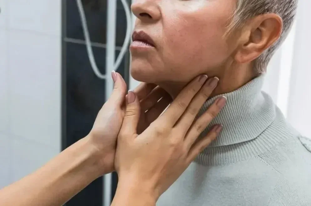 A person with short hair wears a turtleneck sweater while another person's hands gently touch their neck, possibly examining or comforting them.