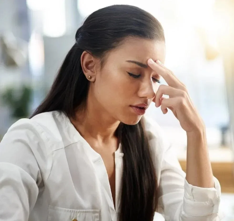 A woman in a white shirt looks stressed, pressing her fingers to her forehead. The setting is a bright, blurred indoor space, conveying tension.
