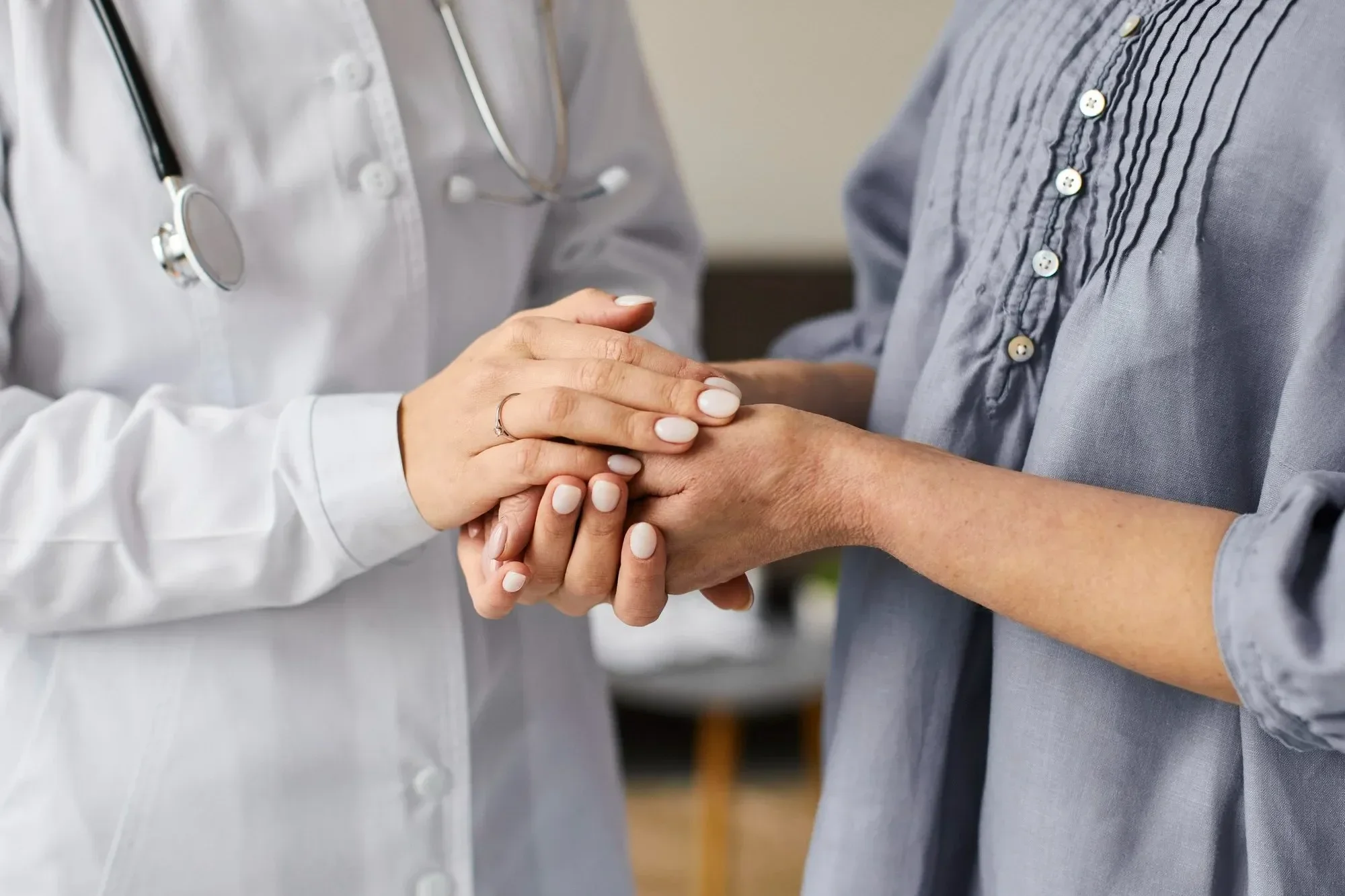A doctor in a white coat holds a patient's hands, offering comfort and support. The scene conveys empathy and care in a medical setting.