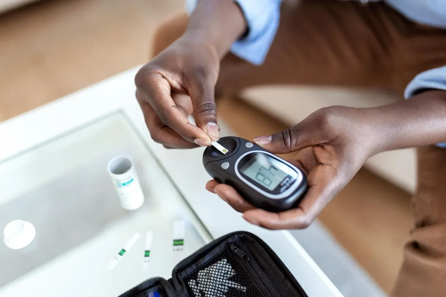 Person using a glucose meter to check blood sugar levels. The device reads 87 mg/dL. Test strips and a container are on a glass table nearby.