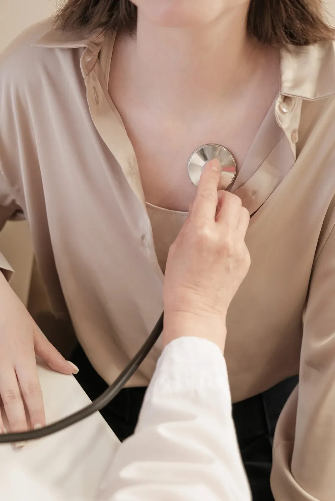 A doctor uses a stethoscope to listen to a patient's heartbeat. The patient wears a beige blouse, and the setting conveys a calm, professional tone.