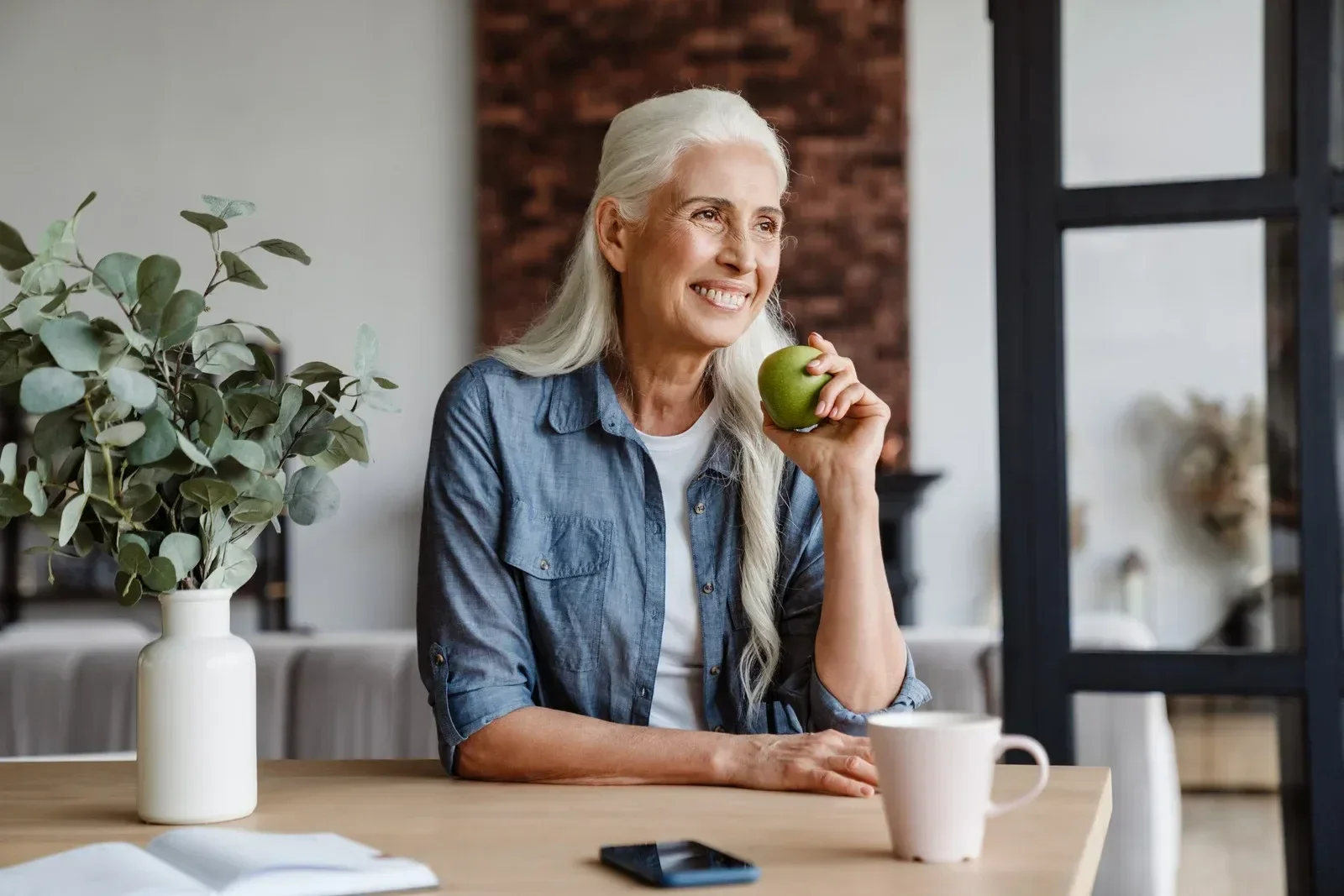 Elderly woman with long gray hair, smiling while holding a green apple. She sits at a table with a vase of leaves, a coffee cup, and a smartphone.