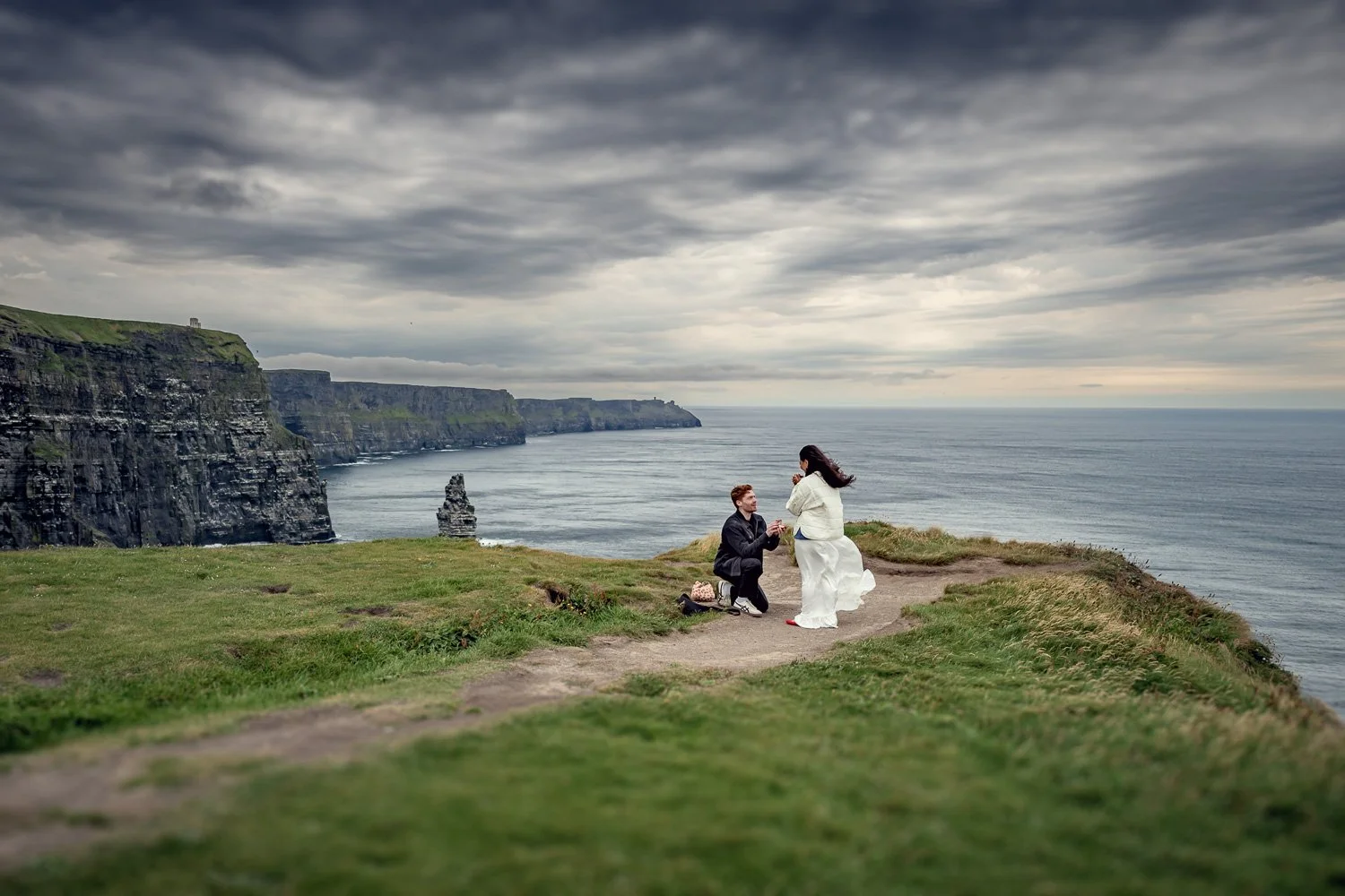 Engagement proposal photography at the Cliffs of Moher capturing the surprise moment as he proposes on a cliffside overlooking the Atlantic Ocean.