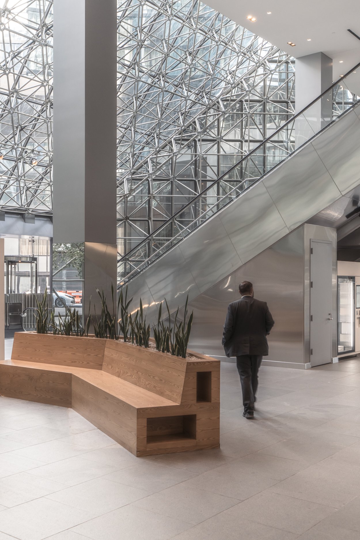 Interior of a modern building with a glass and steel staircase, a man in a suit walking, a wooden bench with plants, and large windows.