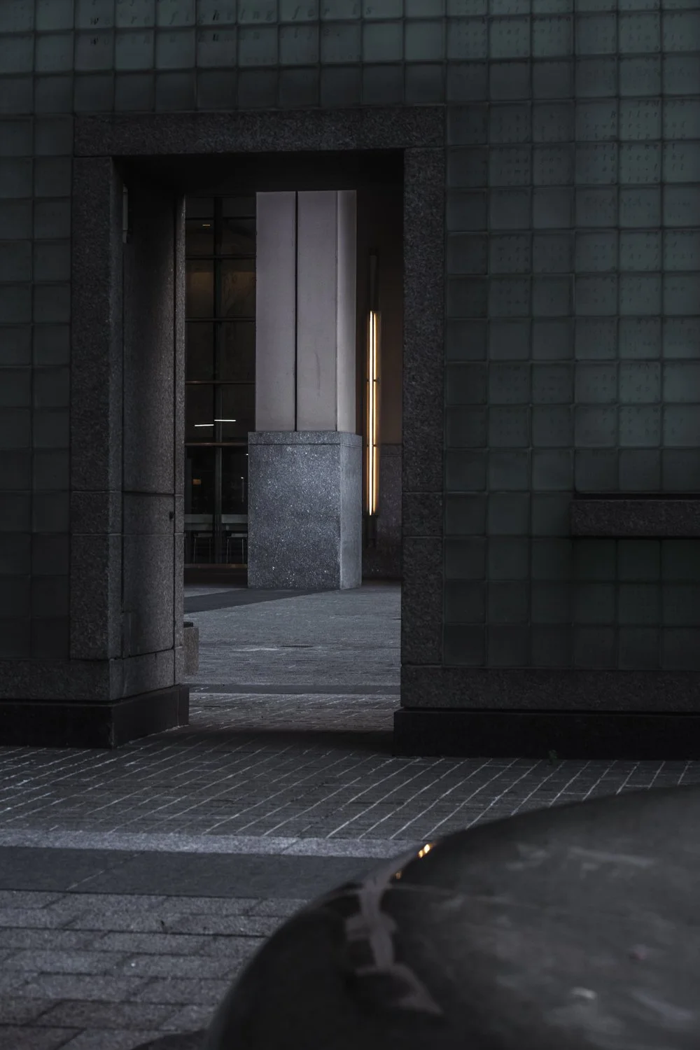 A dark, modern building entrance with a rectangular opening, stone and tile exterior, illuminated vertical light fixtures, and part of a car in the foreground.