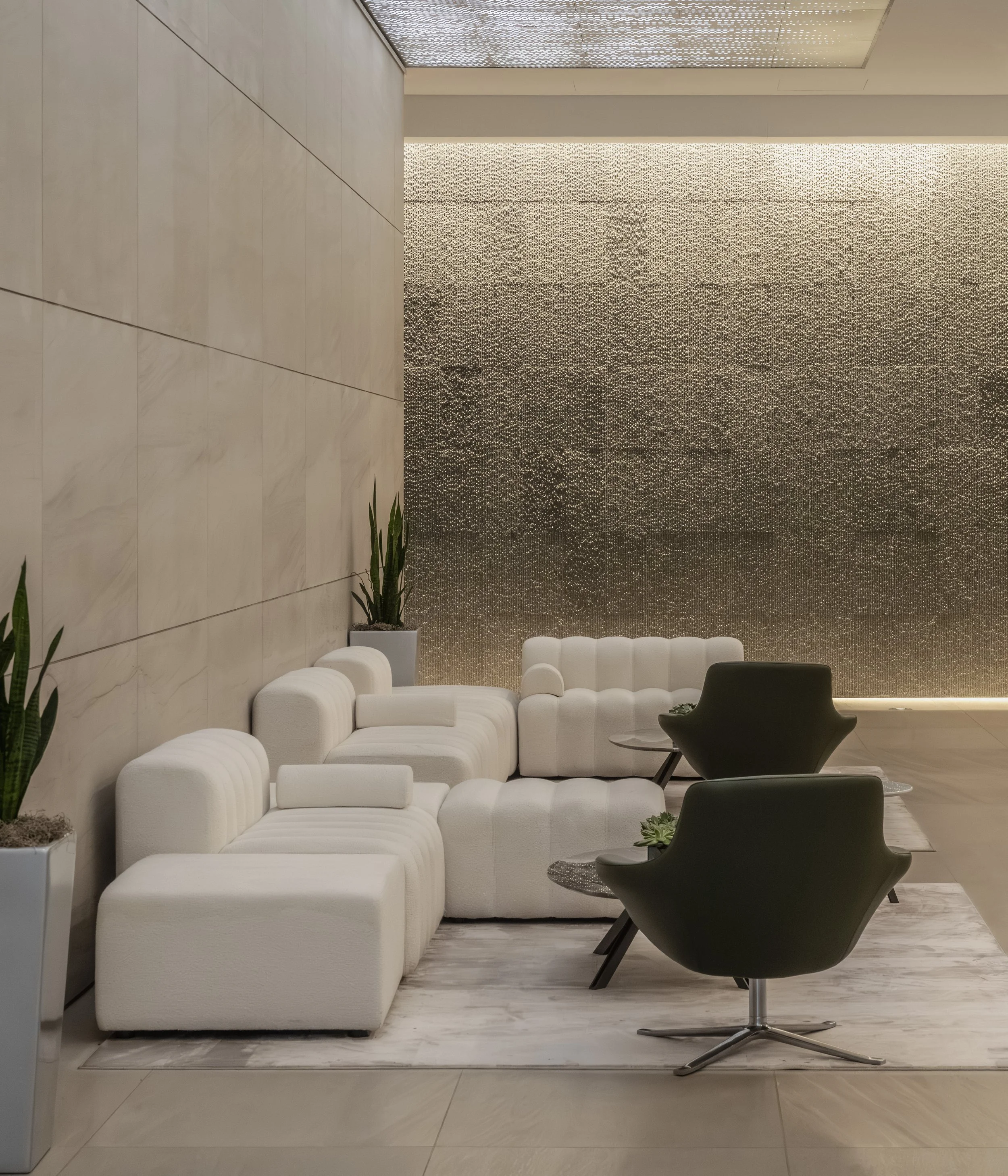 Modern hotel lobby with white seating, black chairs, small side tables, and potted plants, with textured wall backdrop and soft lighting.