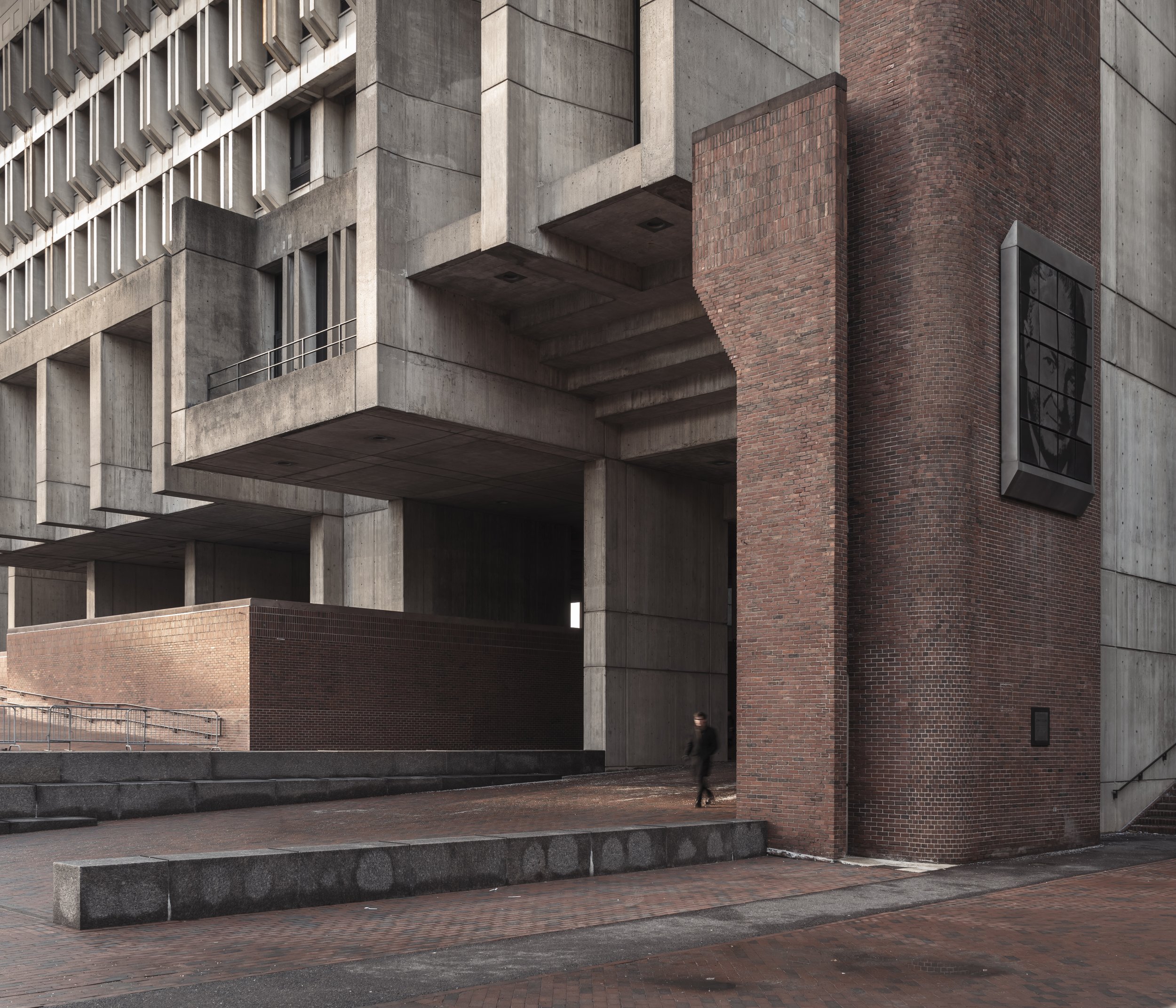 Modern building with concrete and brick architecture, featuring rectangular balconies and stairs, with a person walking on a brick sidewalk.
