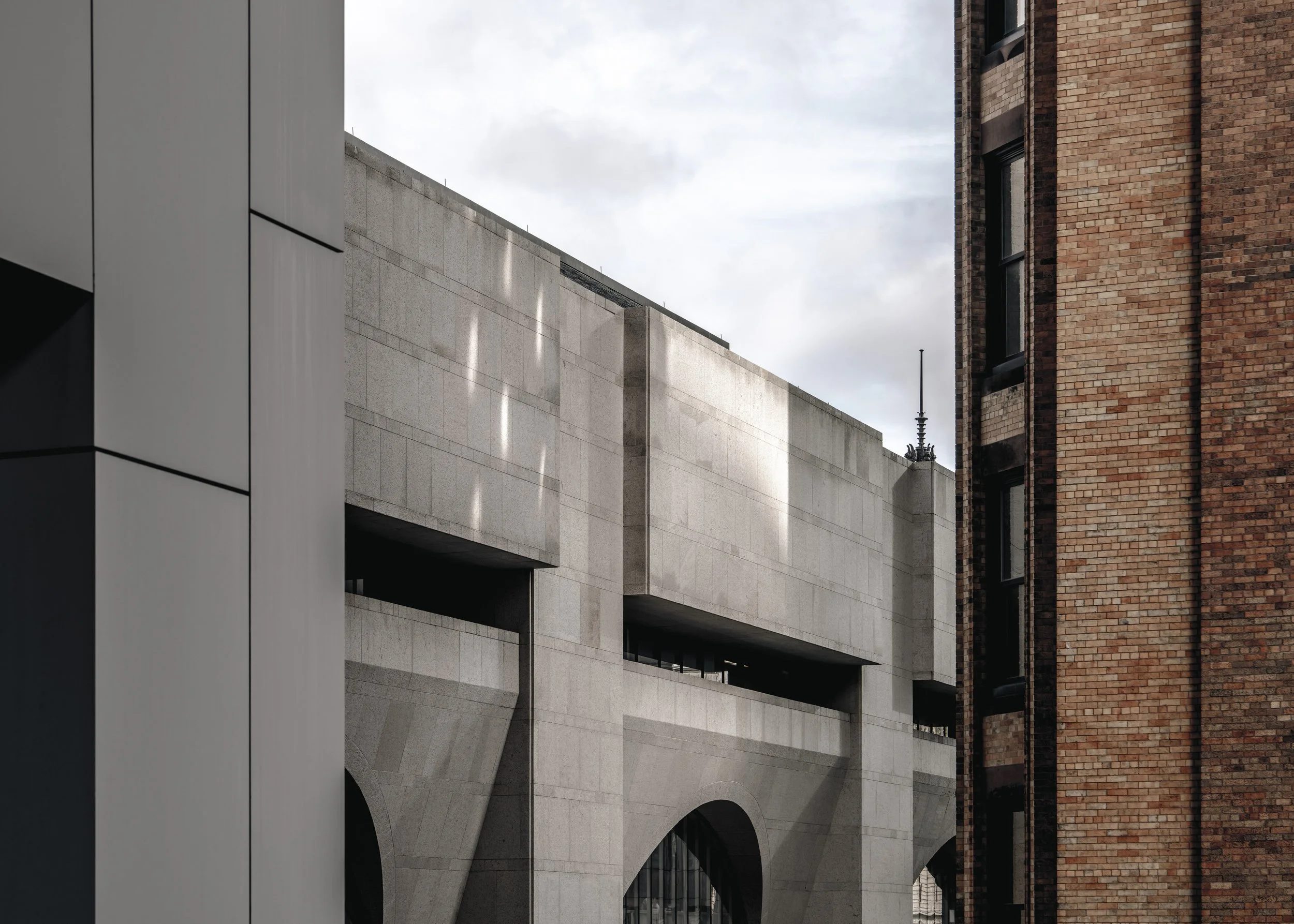 Close-up view of a modern grey concrete building with arches and a red brick building adjacent on the right.