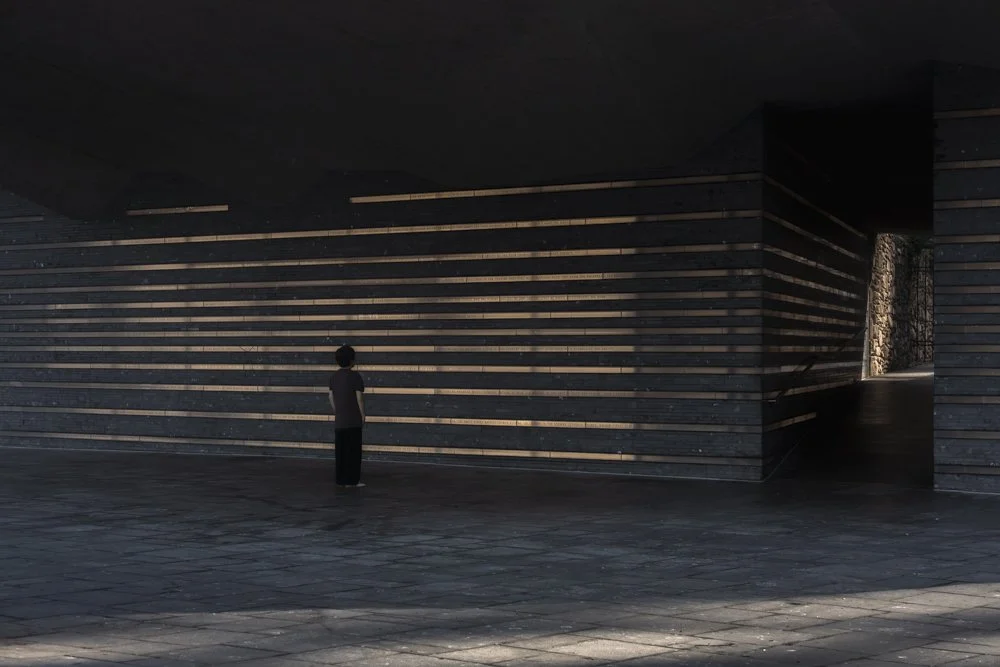 A person standing in front of a modern architectural wall with horizontal wooden slats, casting shadows, and an opening revealing greenery outside.