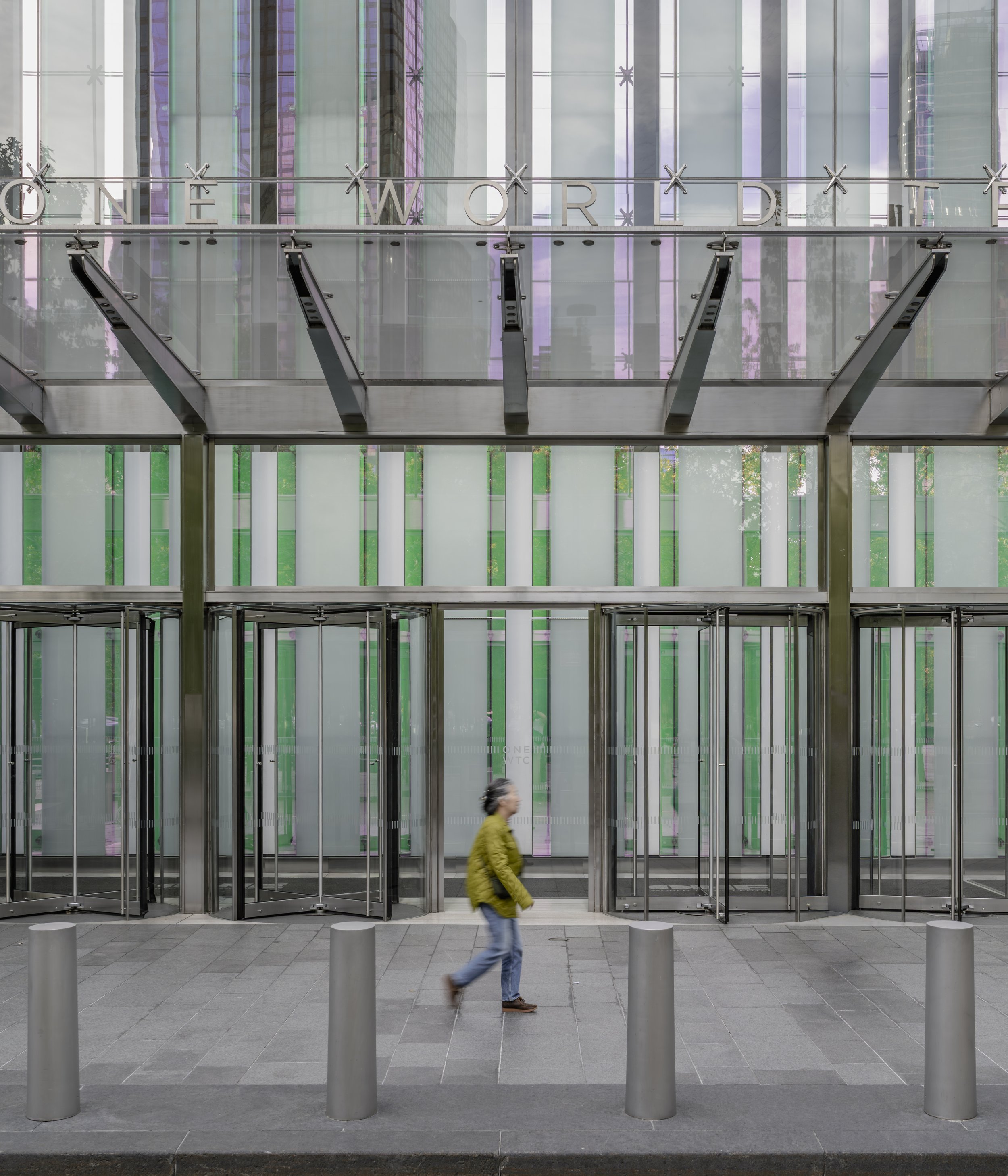 Person walking past the modern entrance of One World Trade Center with glass doors and a green and purple glass facade.