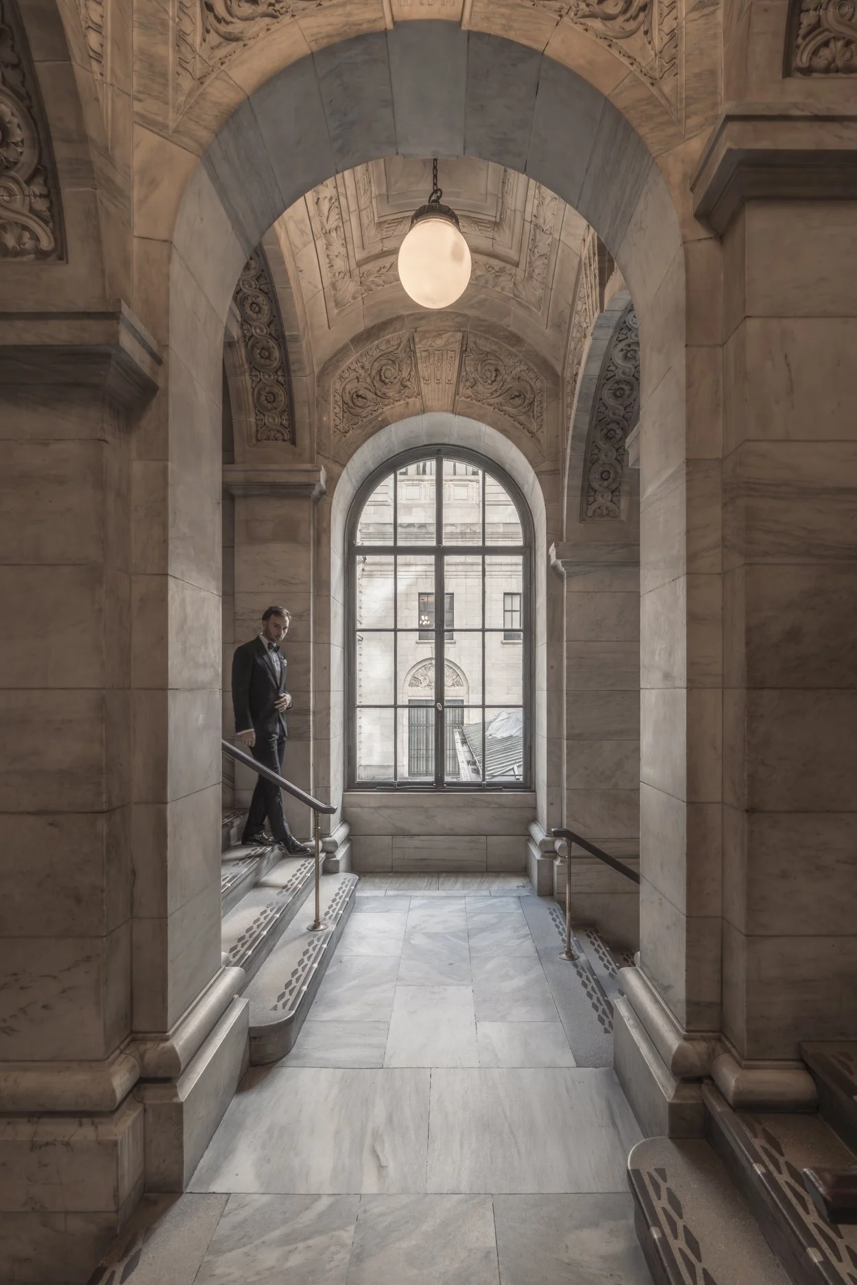 A man in a suit standing on marble stairs in an ornate stone hallway with a large arched window and detailed ceiling carvings.