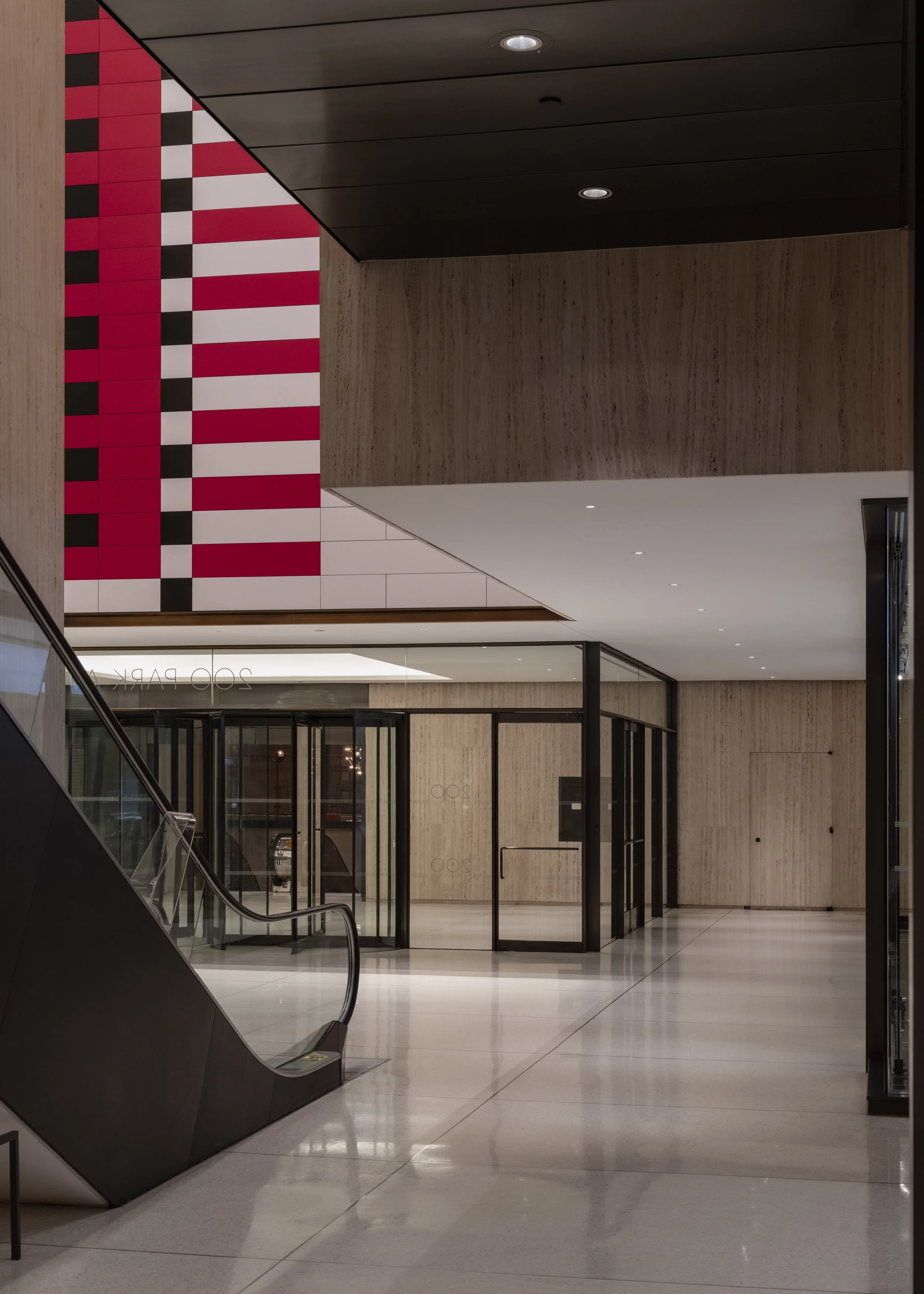 Empty modern lobby with escalator, glass doors, wood paneling, and an abstract red, black, and white artwork on the ceiling.