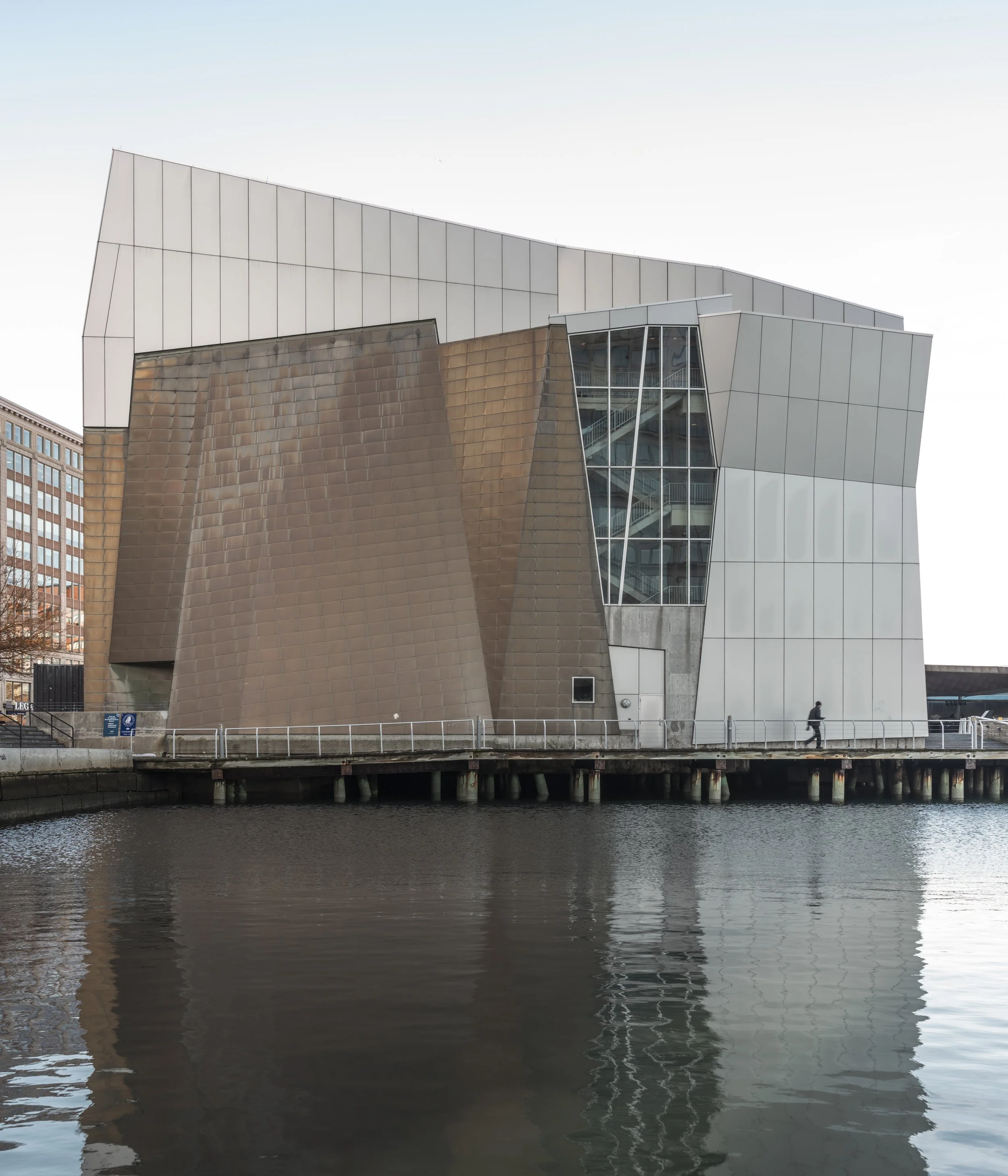 Modern building with unique geometric design by water, featuring brown, gray, and glass panels, with reflections in the water.